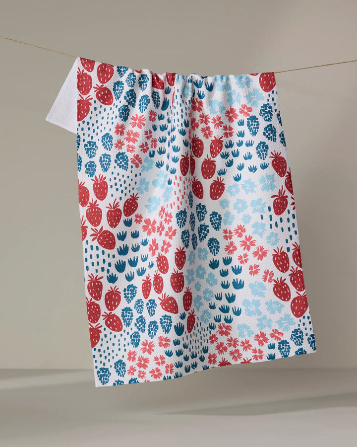 The Berry Meadow kitchen towel by Hufton Studio, made from recycled material, features red strawberries with blue flowers and leaves on white fabric and hangs from a string against a neutral background.