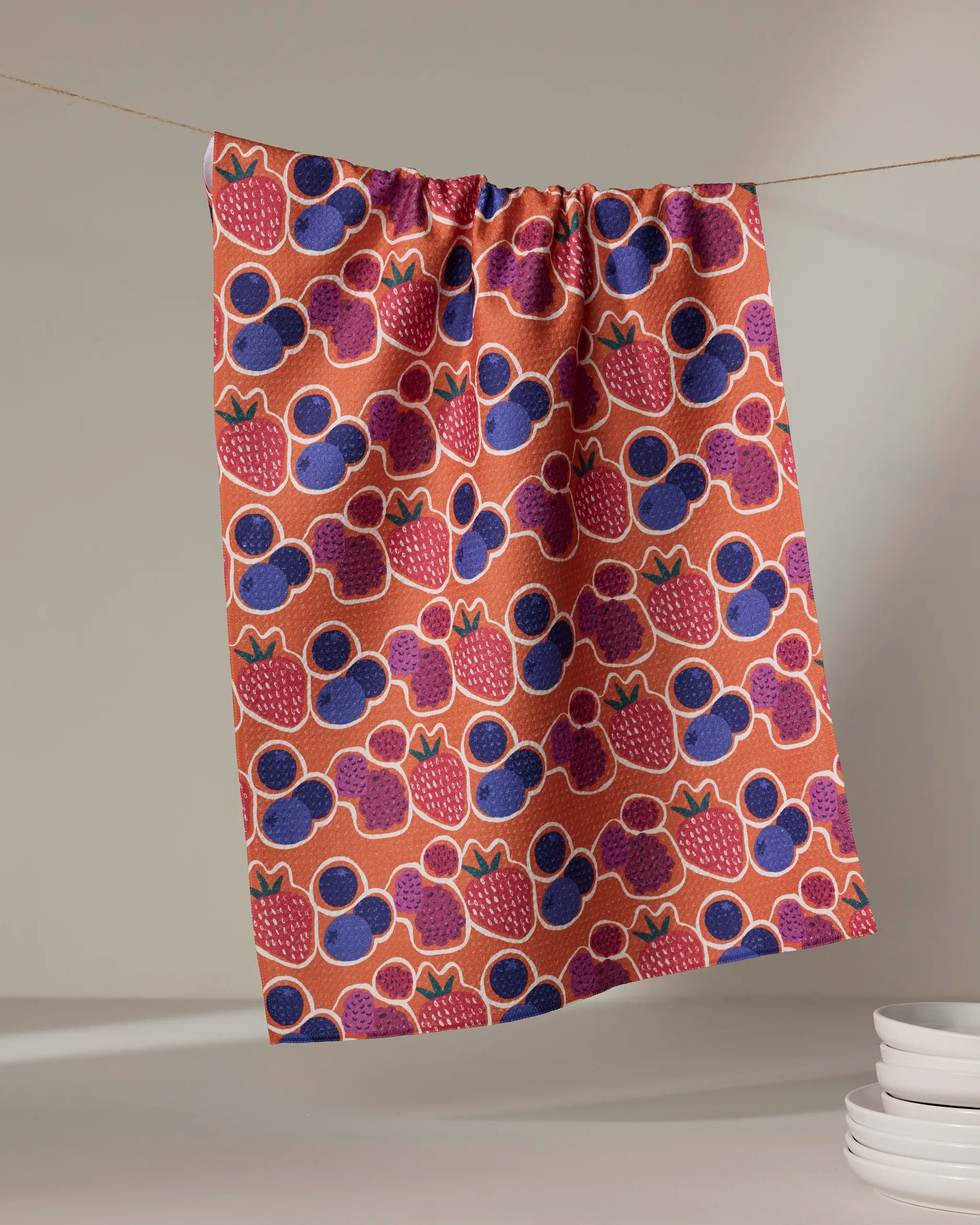 The Berrylicious tea towel by Lizzie Clark, made from recycled materials, features a quick-drying colorful strawberry and blueberry pattern. It hangs against a plain backdrop with white plates stacked in the lower right corner.