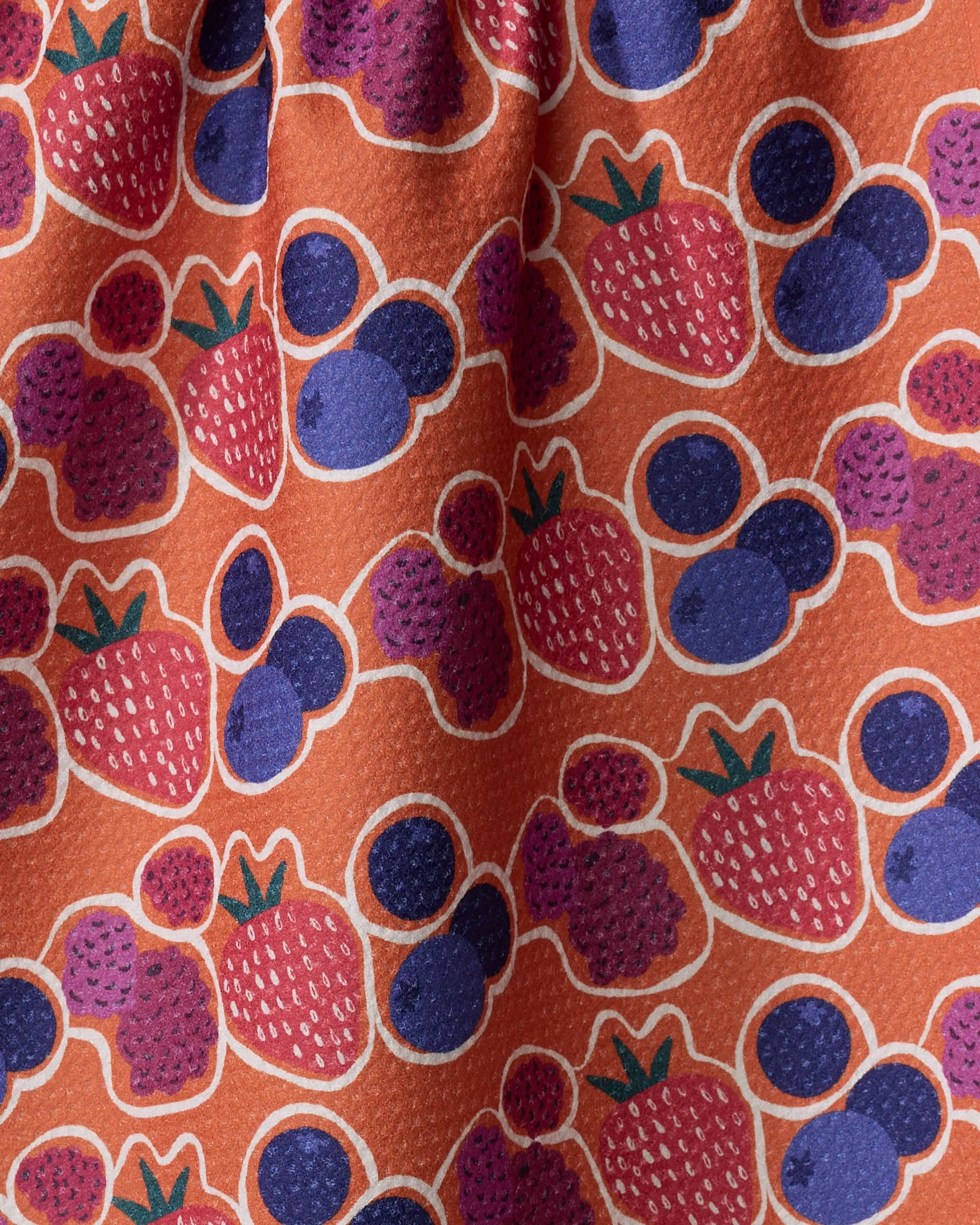 The Lizzie Clark "Berrylicious" tea towel is made from recycled materials and showcases a vibrant orange background with red strawberries and dark purple blueberries in a textured, slightly wrinkled fabric.