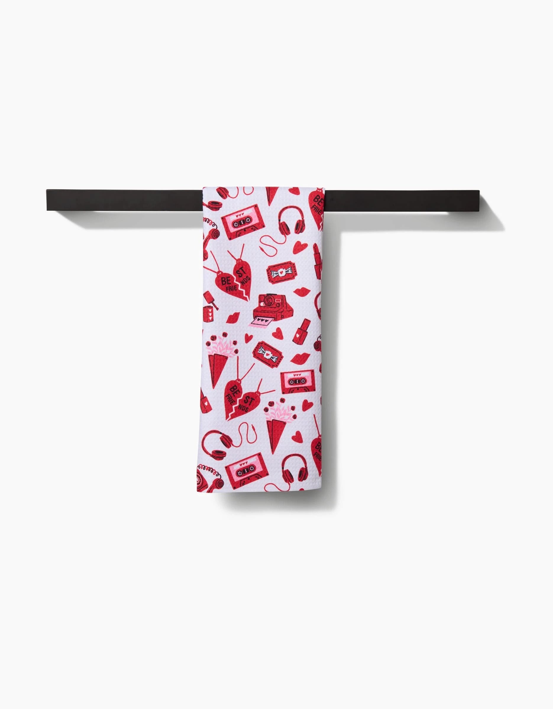 The Best Friends tea towel by Charlie Rowan Designs, featuring red illustrations of retro electronics, hangs neatly on a black bar against a plain white background.