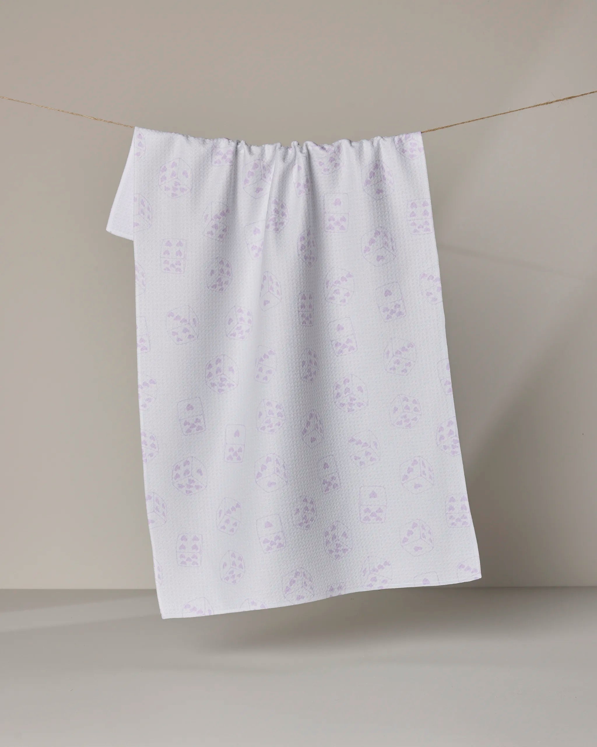 The Bet on You White tea towel by Geometry features a subtle pattern of light purple cat faces on white. Made from recycled materials, it's eco-friendly and stylish—shown hanging on a clothesline against a neutral background.