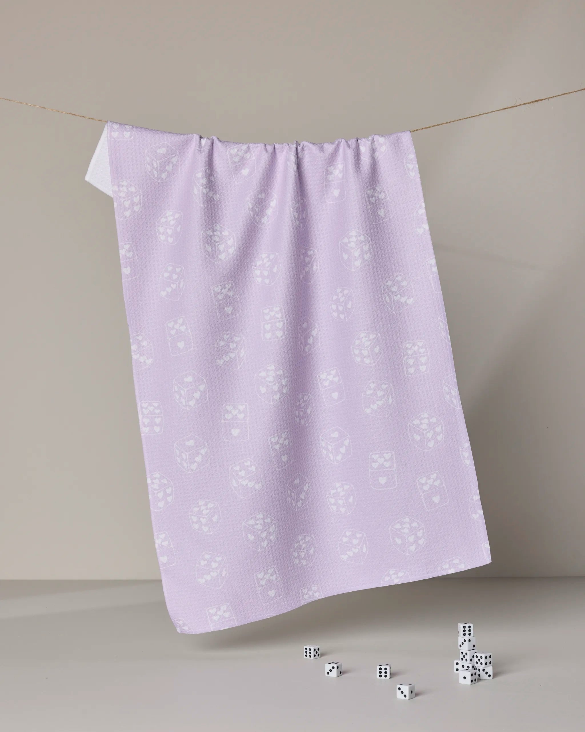 The Geometry "Bet on You" tea towel, featuring an ultra-absorbent pale lavender fabric and white dice pattern, hangs elegantly with matching small white dice below against a neutral backdrop.