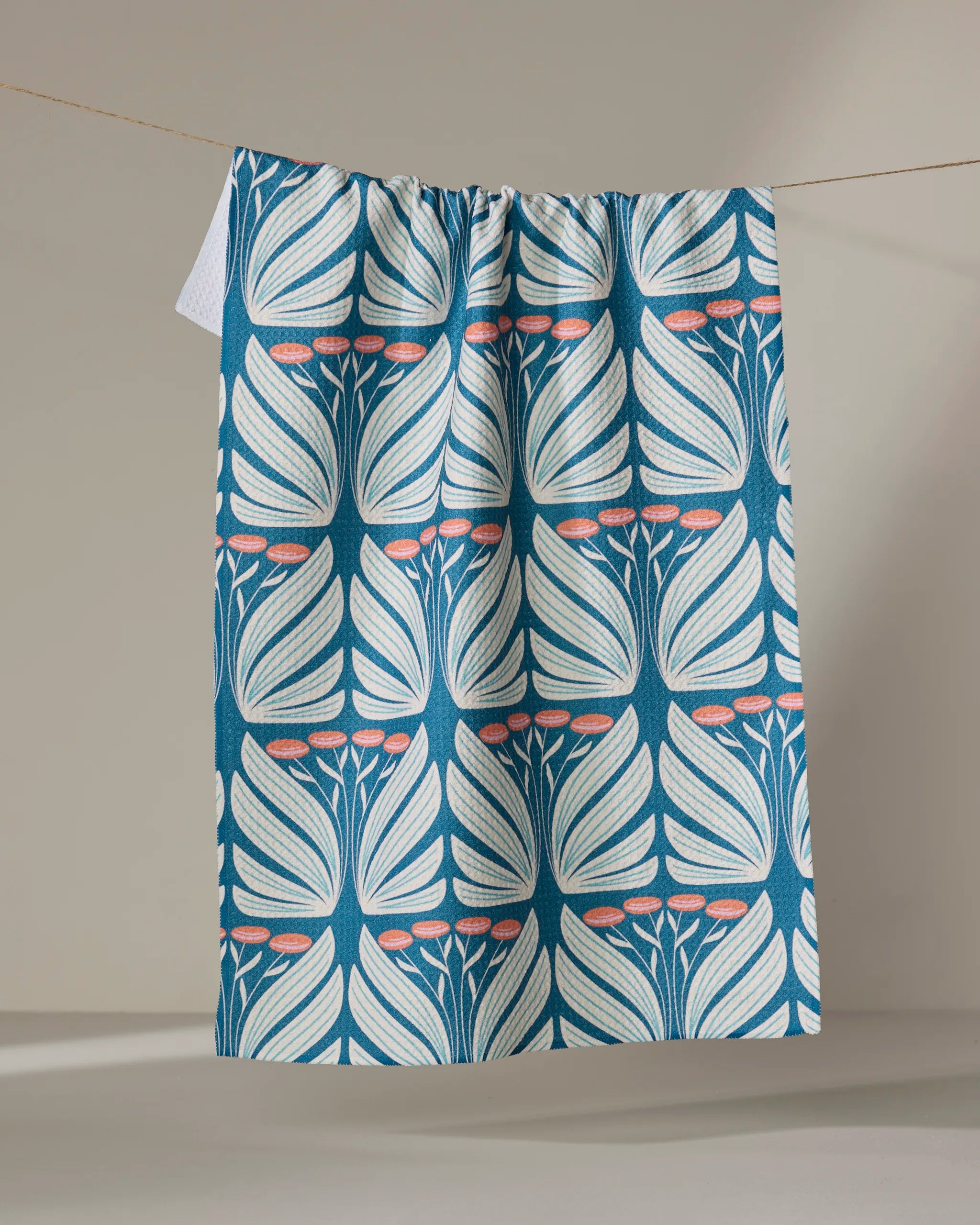 Beyond the Gate" by Rebecca Woolbright—a blue and white tea towel with a symmetrical leaf and pink flower design, made from recycled materials—hangs on a clothesline against a neutral background.
