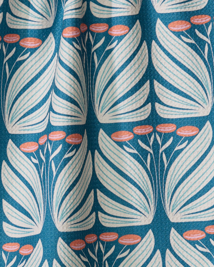 Beyond the Gate by Rebecca Woolbright is a retro botanical tea towel in blue, featuring white stylized leaves and small orange oval flowers. It’s made from quick-drying recycled material and softly gathered for an elegant look.