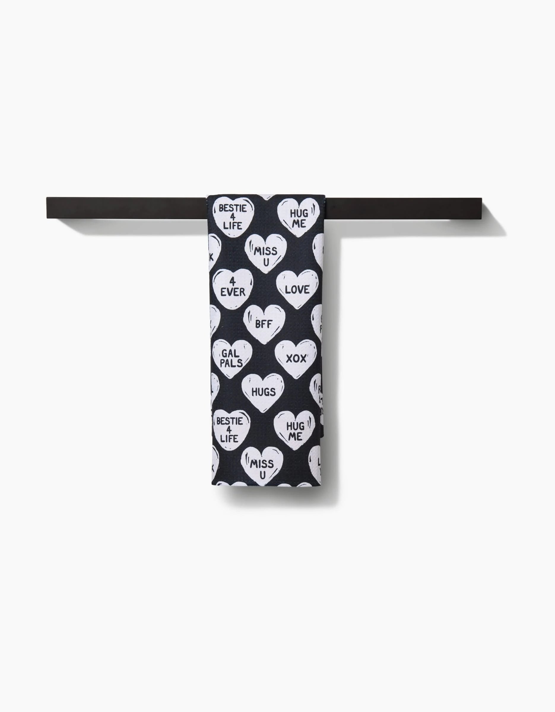 The BFF Candy Hearts towel by Hufton Studio—a black, quick-drying towel with white heart patterns and phrases—hangs neatly over a simple black bar against a white background.