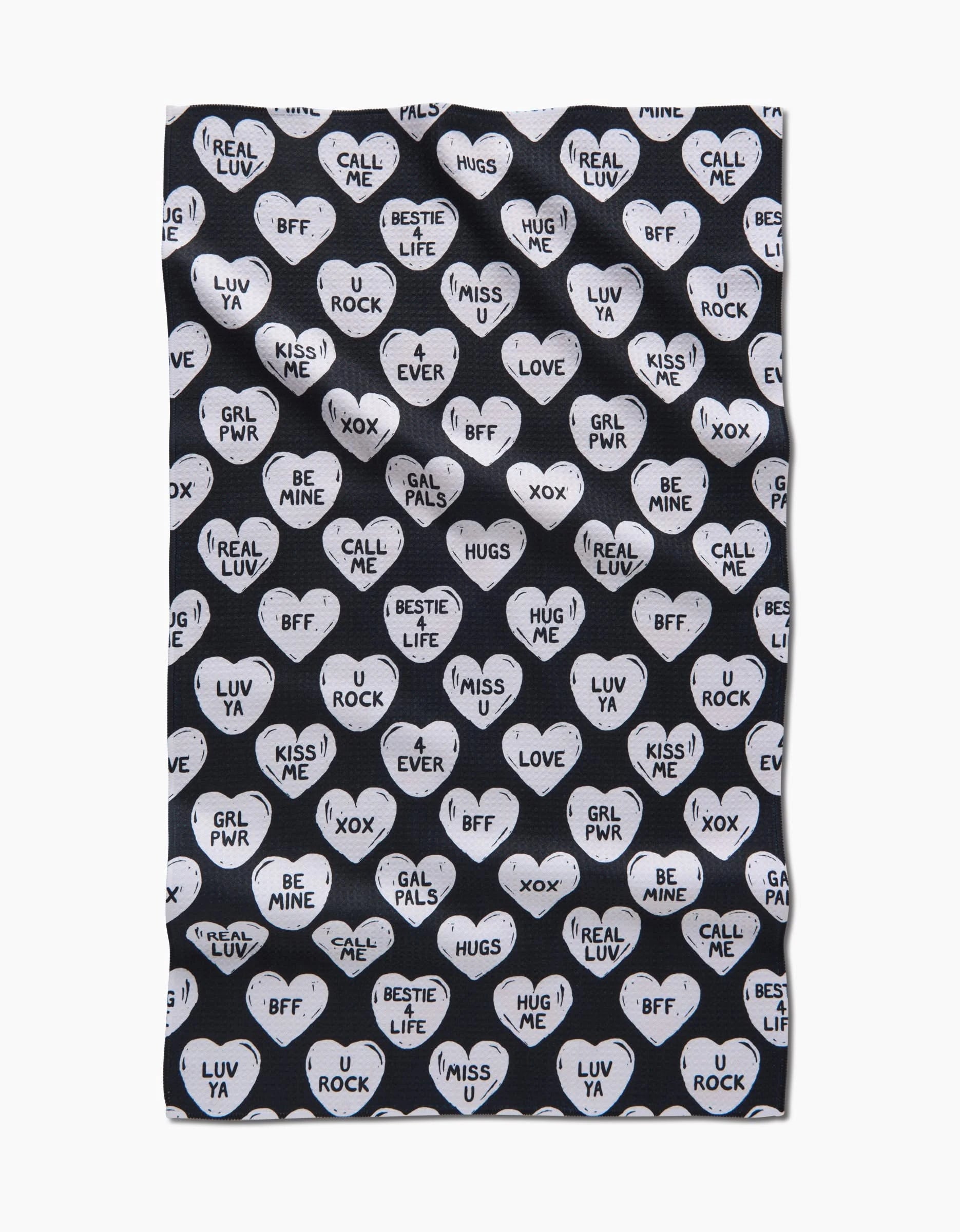 The BFF Candy Hearts tea towel by Hufton Studio features a repeating pattern of white heart-shaped candies with black text messages such as GRL PWR, BFF, LUV YA, MISS ME, XOX, and U ROCK on a bold black background.
