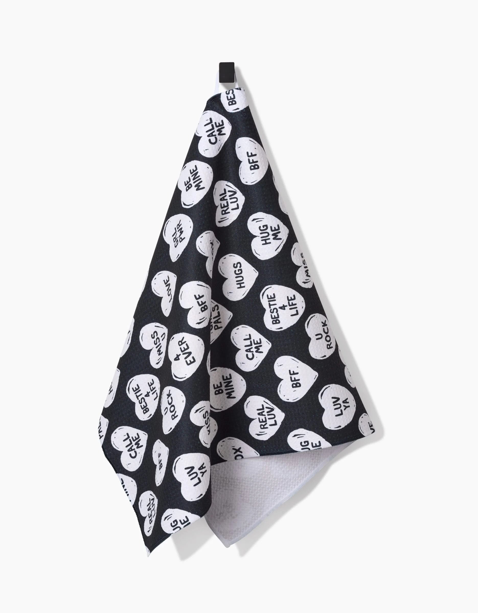 The Hufton Studio BFF Candy Hearts kitchen towel, made from recycled materials, features white heart-shaped designs with phrases like HUG ME and CUTIE. The black towel is displayed hanging on a hook against a white background.