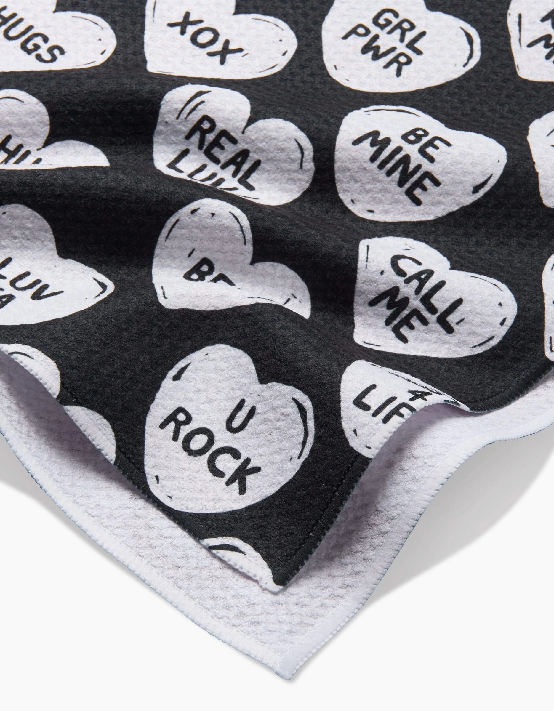 The BFF Candy Hearts kitchen towel by Hufton Studio is black and white, made from recycled fabric, and features heart-shaped candies with messages like XOX, GRL PWR, BE MINE, CALL ME, U ROCK, and HUGS. One fabric corner is curled up.