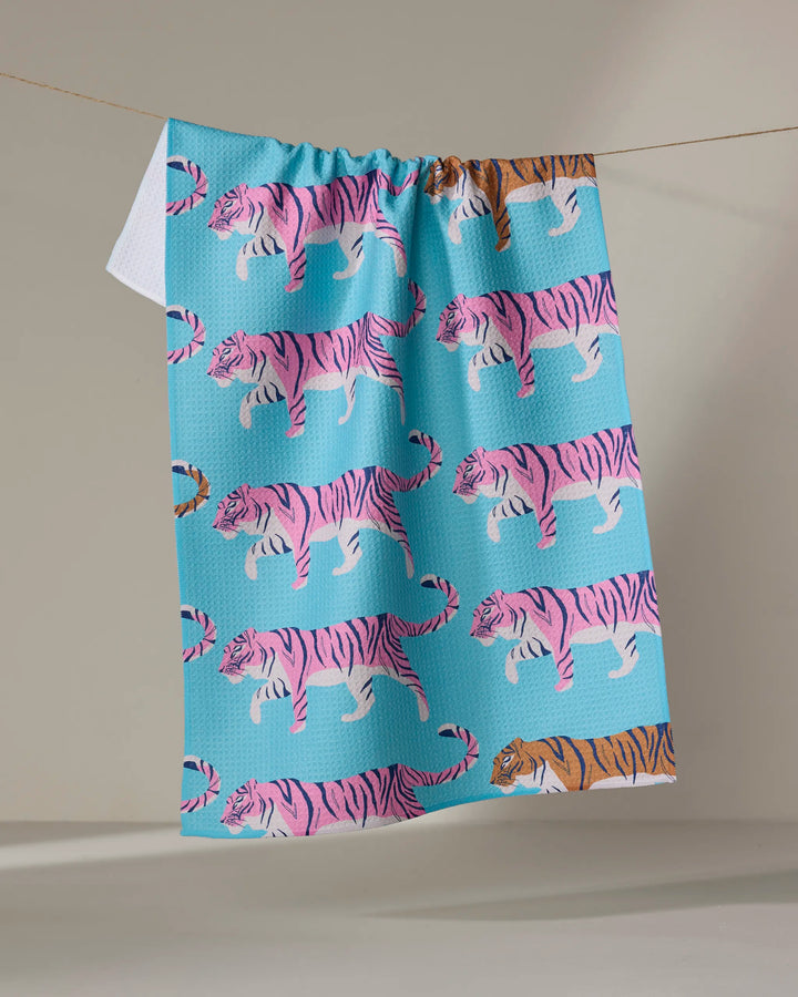 The "Big Cats" tea towel by Julianne Haness, made from recycled materials, features pink tigers with one orange tiger near the bottom and hangs over a clothesline against a neutral background.