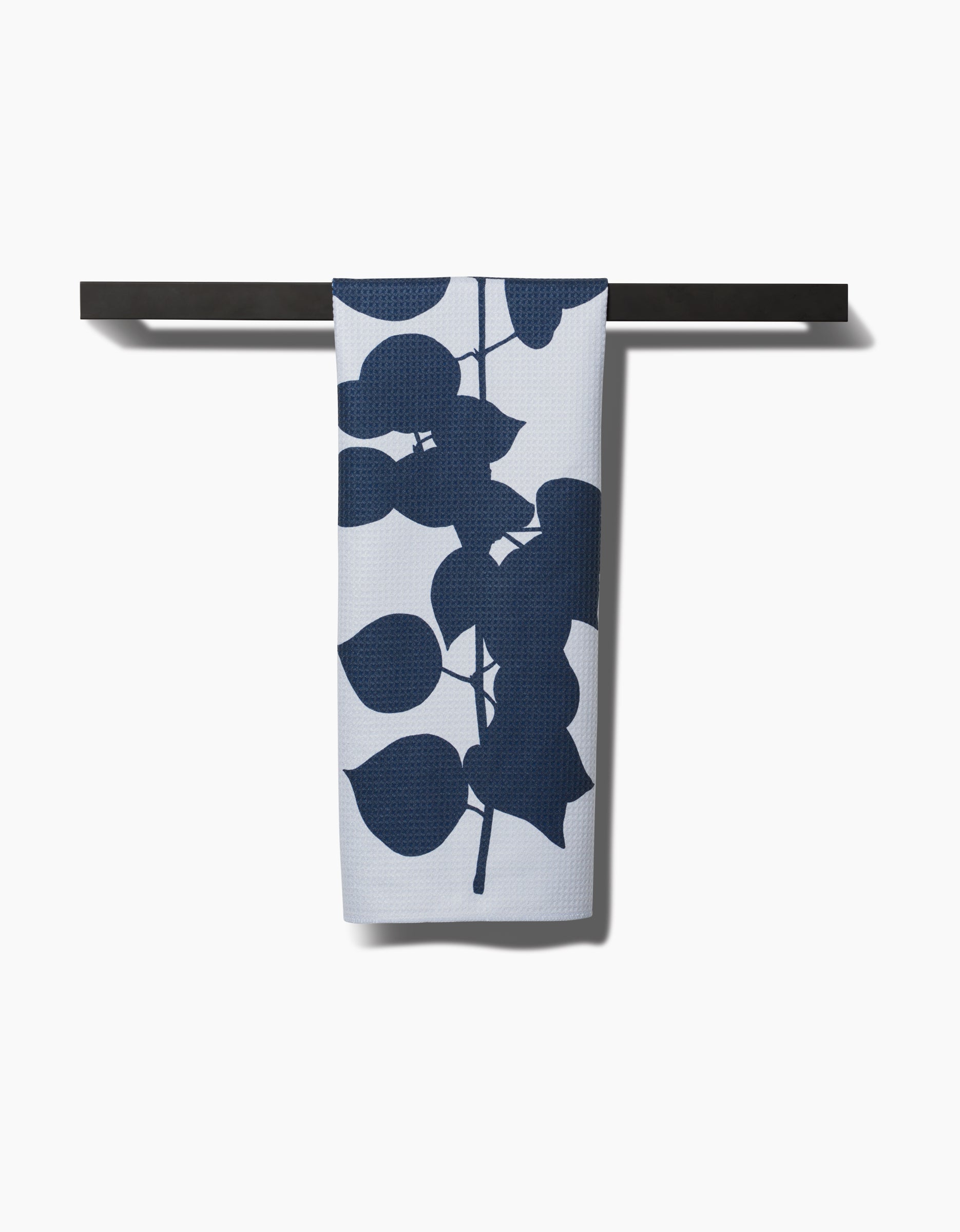 The Birch tea towel by PrintedPlant, featuring a bold dark blue leaf design, hangs neatly on a black towel bar against a plain white background.