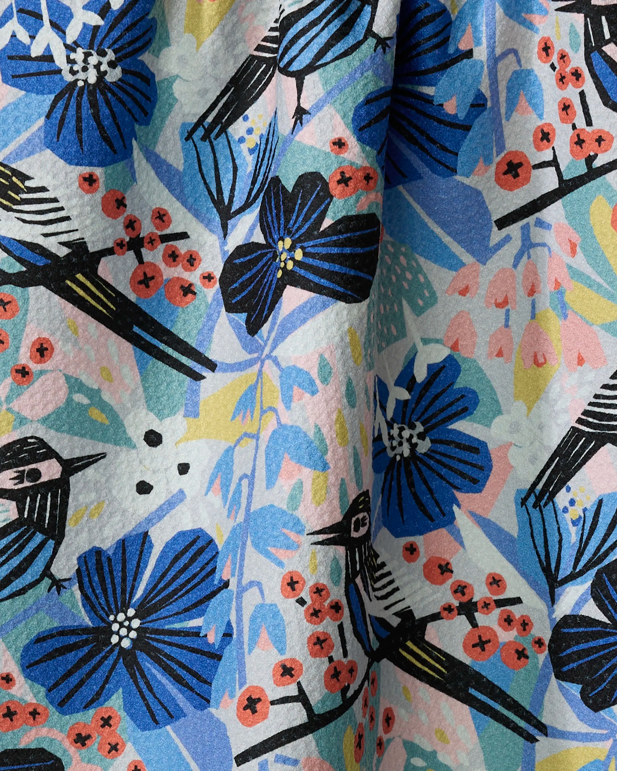 The "Birds And Berries" tea towel by Masha Volnova is made from recycled materials and features a colorful design with blue flowers, red berries, and stylized birds with black and white wings among vibrant abstract shapes.
