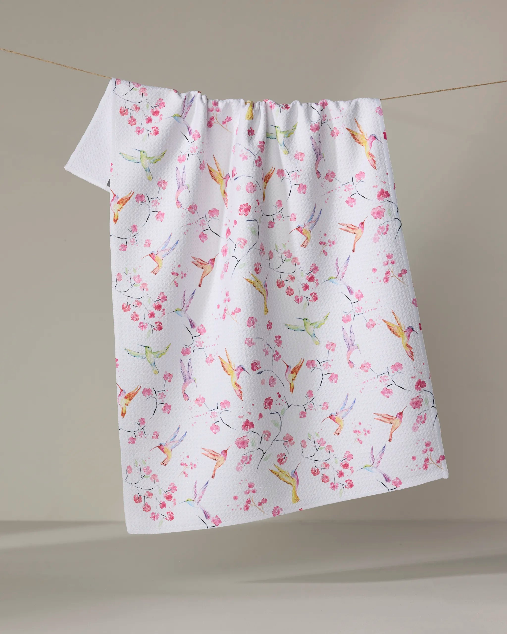 The Elena Fay "Birds and Blooms" tea towel is white, quick-drying, and textured. Hanging on a clothesline, it showcases colorful birds and pink cherry blossoms on a cheerful, delicate background that's plain and neutral.