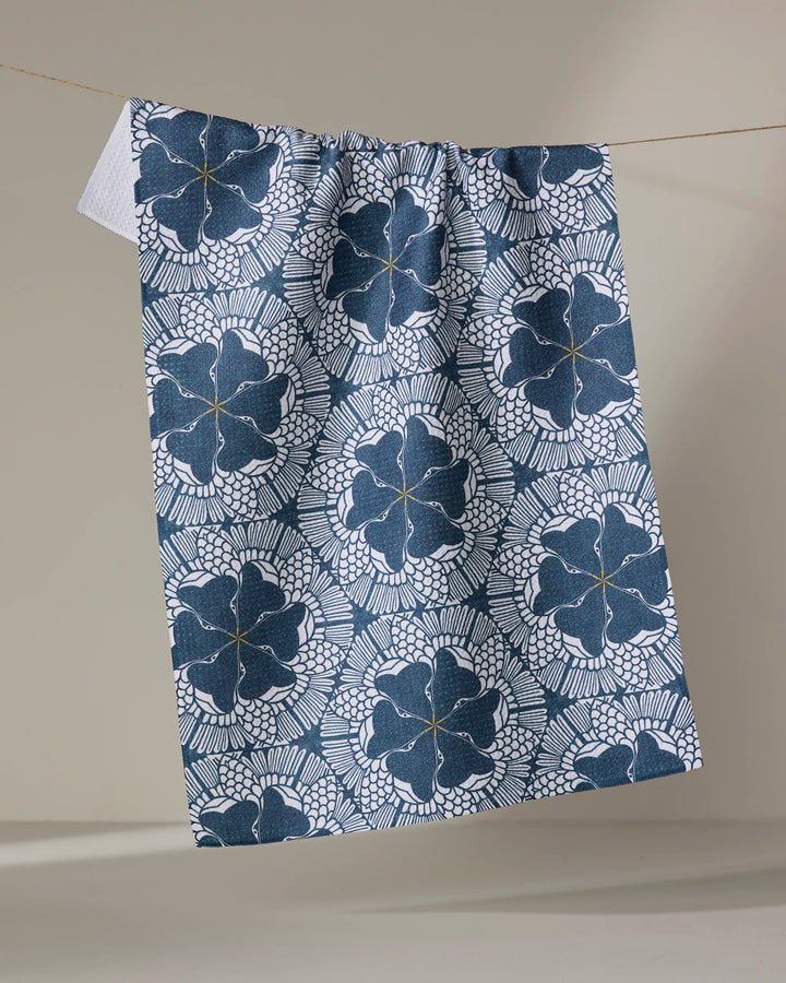 The "Birds of a Feather" tea towel by Renee Fly, featuring blue and white floral and geometric patterns and made from recycled materials, hangs on a string against a neutral background.