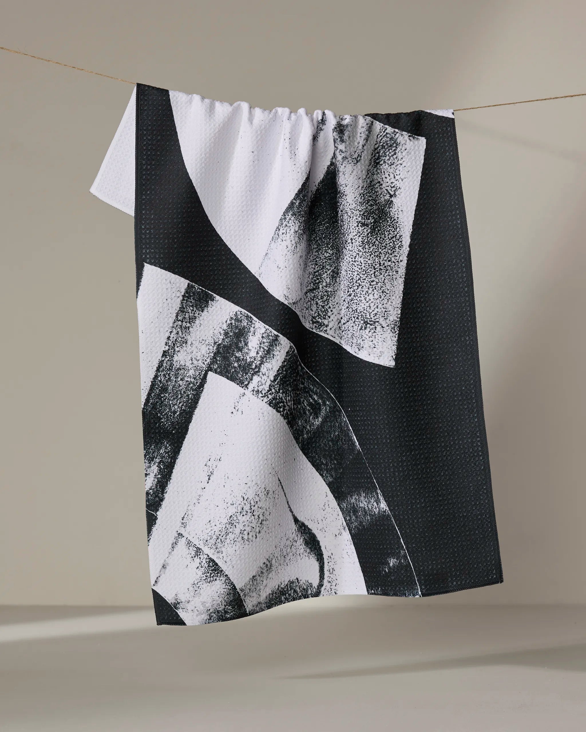 The Skye Walker Black Graphite Shift kitchen tea towel, with black and white abstract geometric patterns, hangs from a string against a neutral backdrop. This ultra-absorbent towel features textured, brush-like markings.