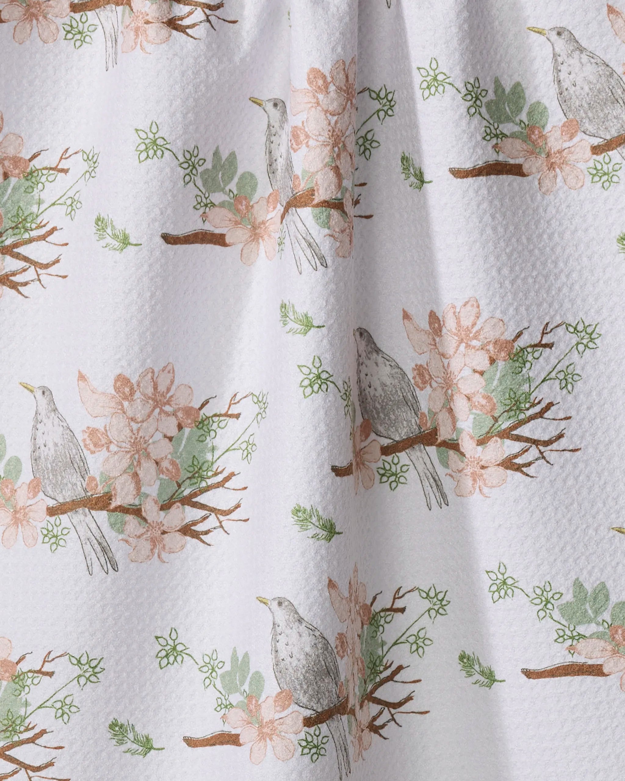 The Blackbird and Blossom tea towel by Linda Fox features gray birds on brown branches with pink flowers and green leaves. This white textured, quick-drying towel offers a soft, nature-inspired look with its repeating printed design.