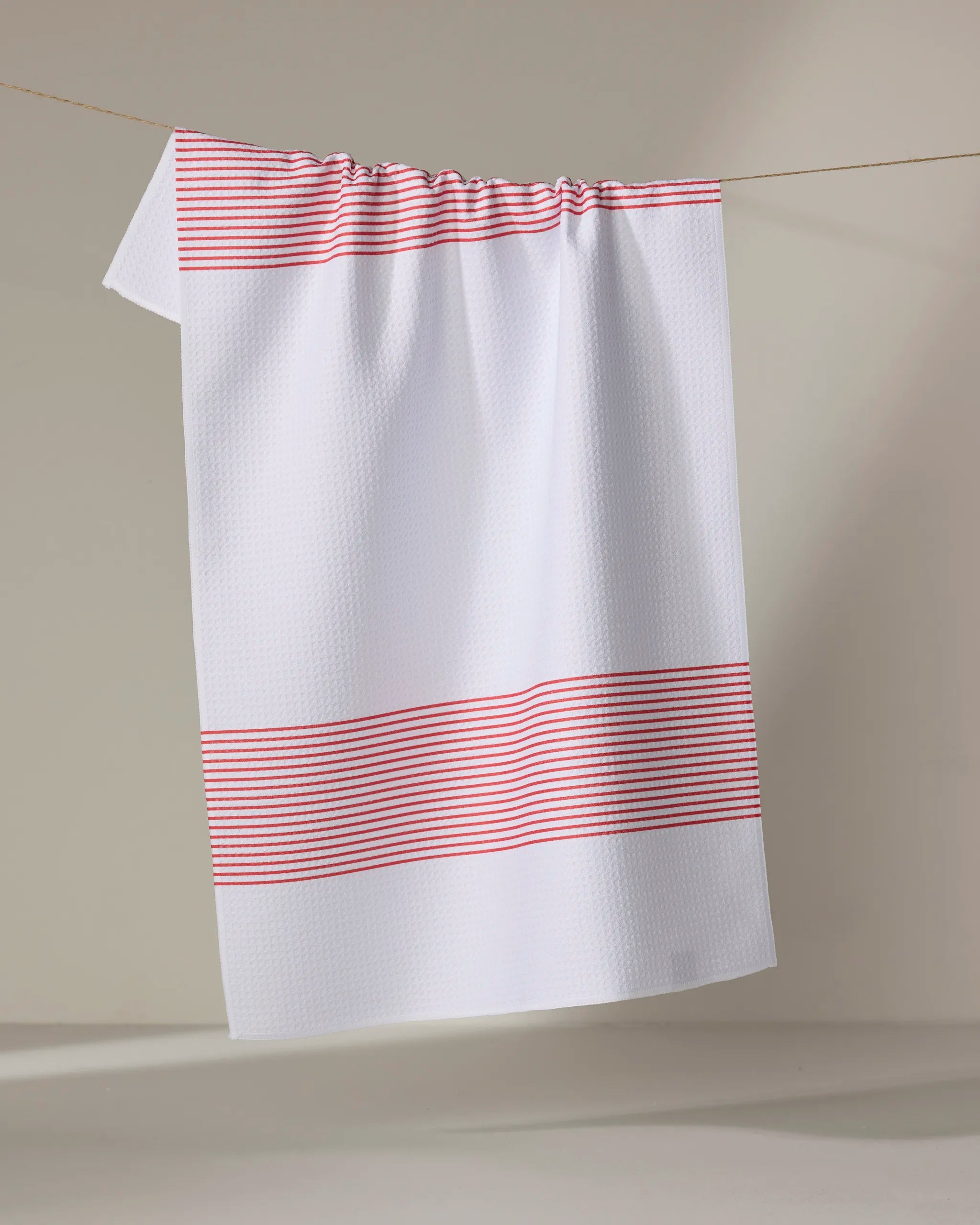 The Buy Guide’s Blank Space - Candy Apple tea towel, made from recycled materials, features a white textured design with red stripes at the top and bottom and hangs on a clothesline against a neutral background.