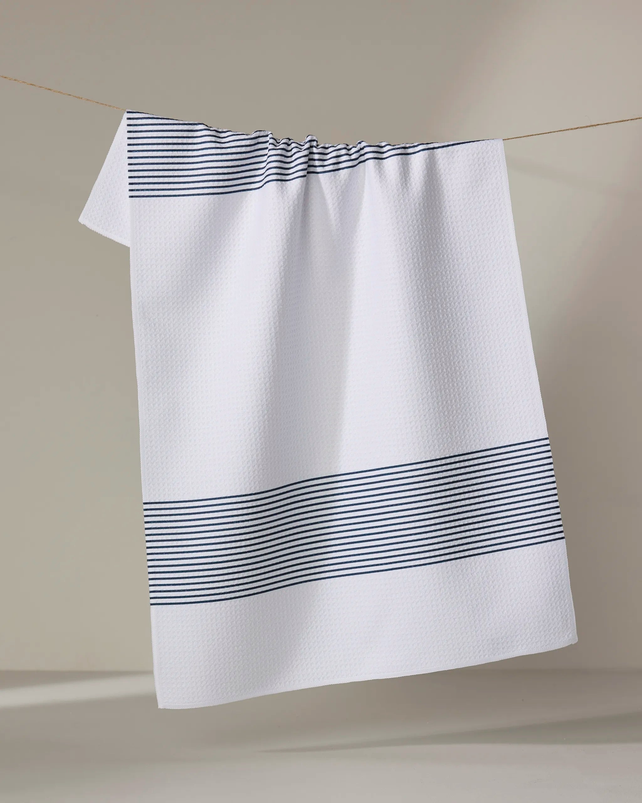 The Buy Guide’s Blank Space - Navy tea towel, crafted from ultra-absorbent material, features a white textured design with horizontal navy stripes near the edges and hangs neatly over a clothesline against a neutral backdrop.
