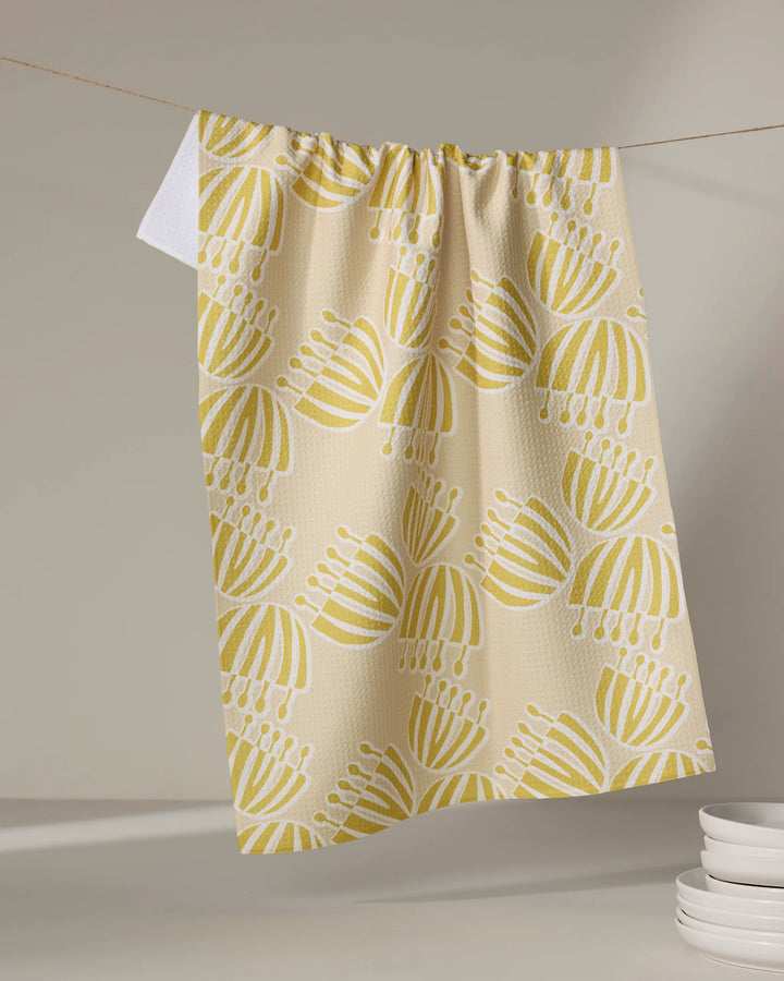The Block Print Tulips tea towel by Lizzie Clark, crafted from recycled materials in a beige with yellow and white tulip pattern, hangs on a string. A white towel and stacked plates appear softly in the neutral background.
