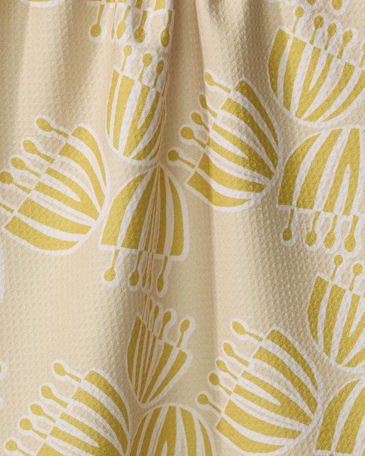 A close-up of the Lizzie Clark Block Print Tulips tea towel, crafted from recycled materials, shows a beige textured surface with a yellow and white abstract tulip pattern of clustered, striped petal shapes with circular ends.
