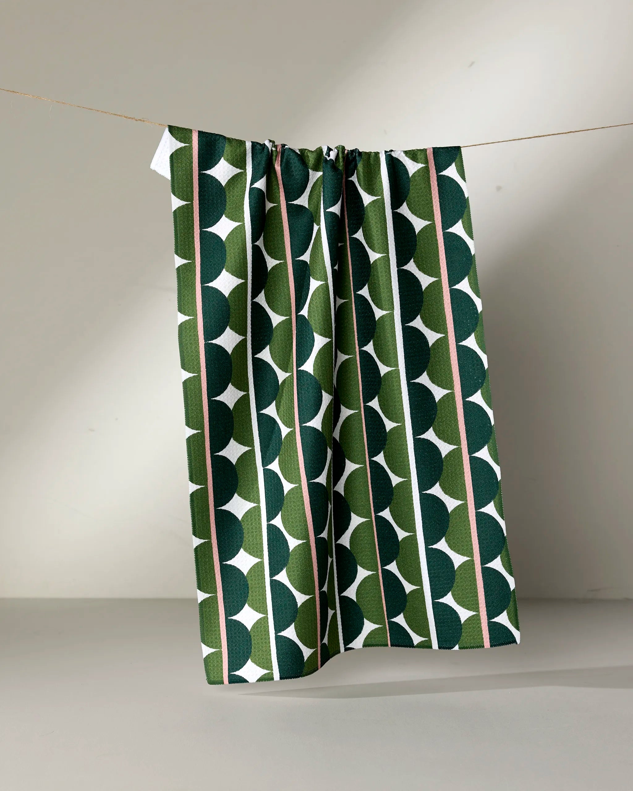 The Rachel Parker Blockprint Eclipse is an eco-friendly kitchen towel featuring green and white geometric patterns and vertical stripes, hanging on a thin string against a plain light background.