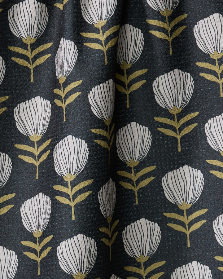 The Blomma tea towel by Anna Christian features a black fabric with white, stylized flowers and gold stems in vertical rows. Ultra-absorbent, it adds elegance and functionality to your kitchen.
