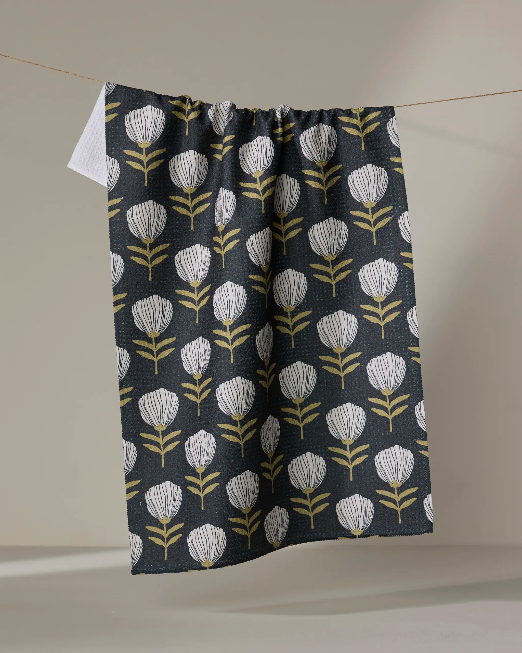 The Anna Christian Blomma tea towel, featuring a dark background and a repeating pattern of white and yellow stylized flowers, hangs on a clothesline against a plain backdrop.