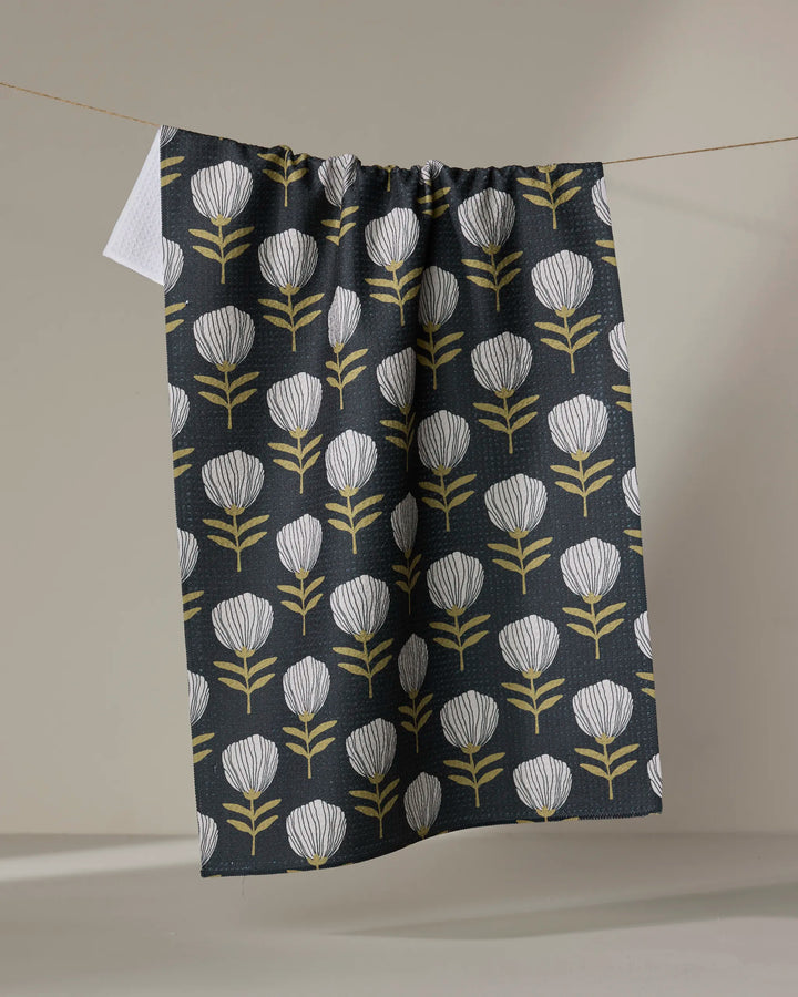 The Anna Christian Blomma tea towel, featuring a dark background and a repeating pattern of white and yellow stylized flowers, hangs on a clothesline against a plain backdrop.