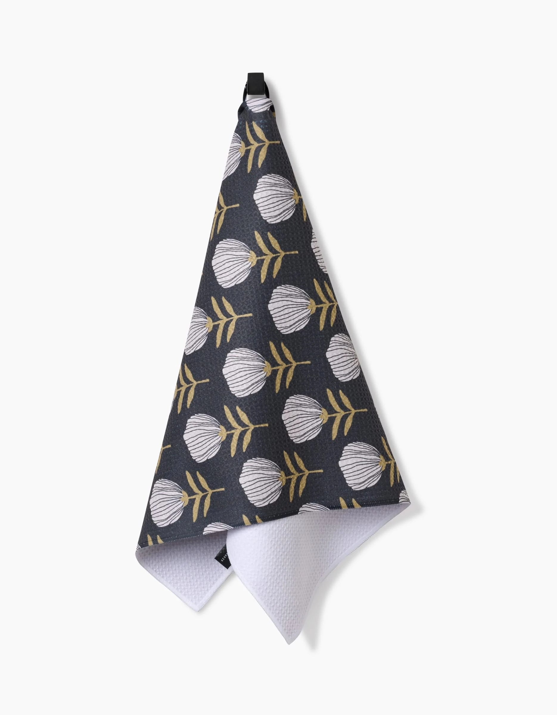 The Blomma tea towel by Anna Christian is made from recycled fibers. It features white flowers with yellow stems on a dark background and hangs from a black loop, displayed against a white backdrop.
