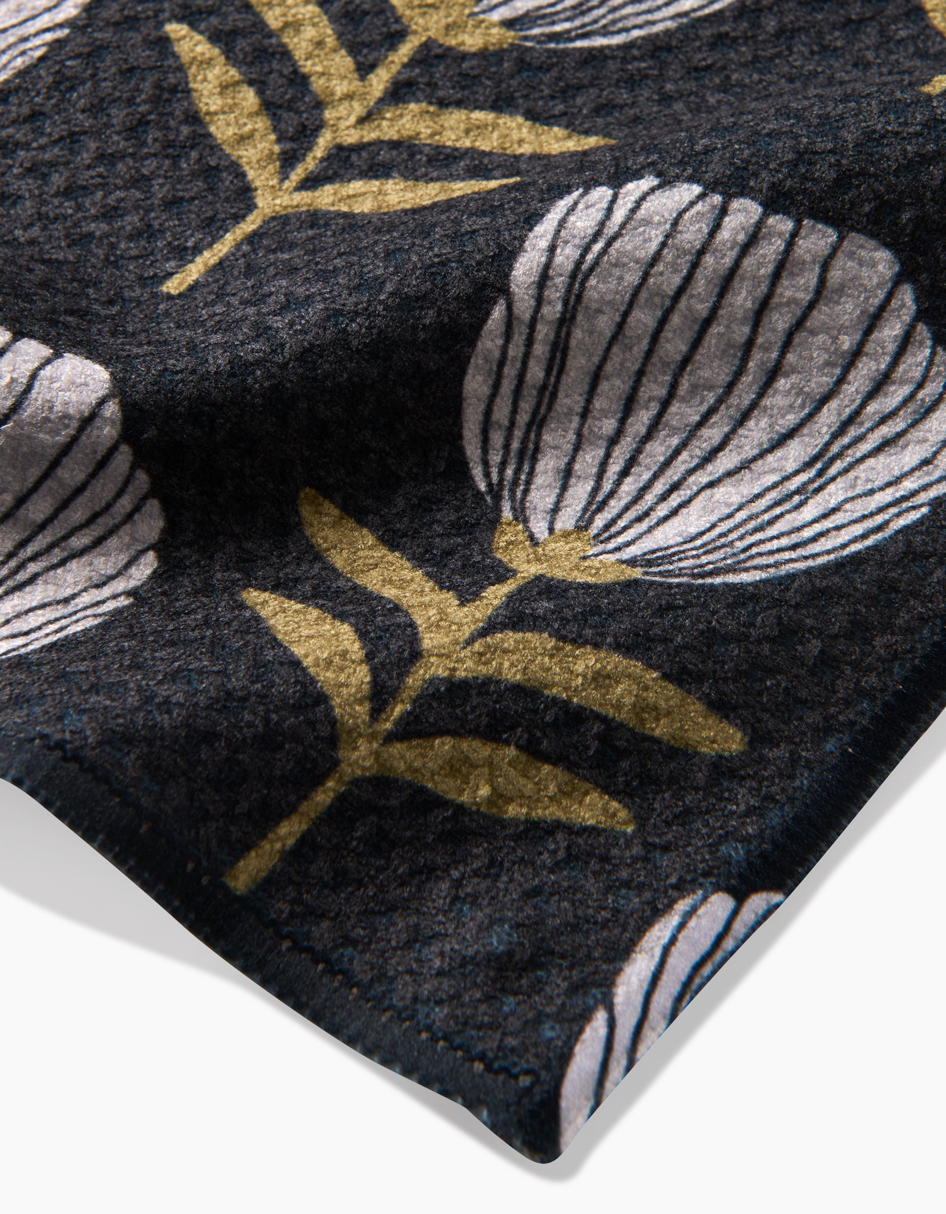 A close-up of the Anna Christian Blomma tea towel shows its dark Geoweave fabric with a floral pattern of white flowers outlined in black and gold leaves, plus visible stitching along the edge.