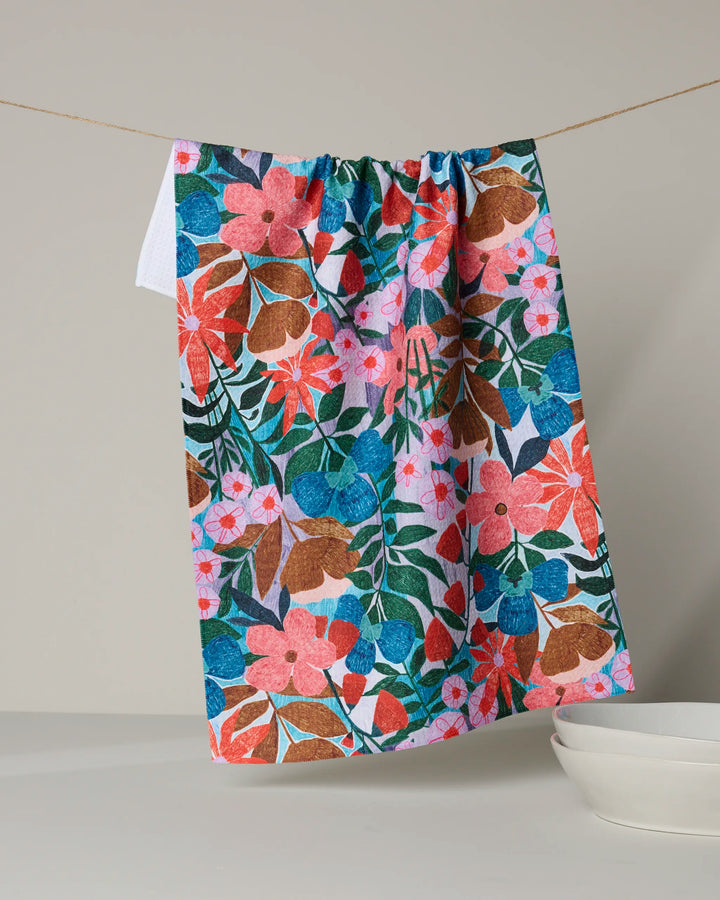 Bloom Entangled by Lisa Nelson is a vibrant, eco-friendly tea towel featuring pink, blue, green, and brown floral patterns. Made from recycled materials, it hangs above white dishes for a colorful touch in your kitchen.