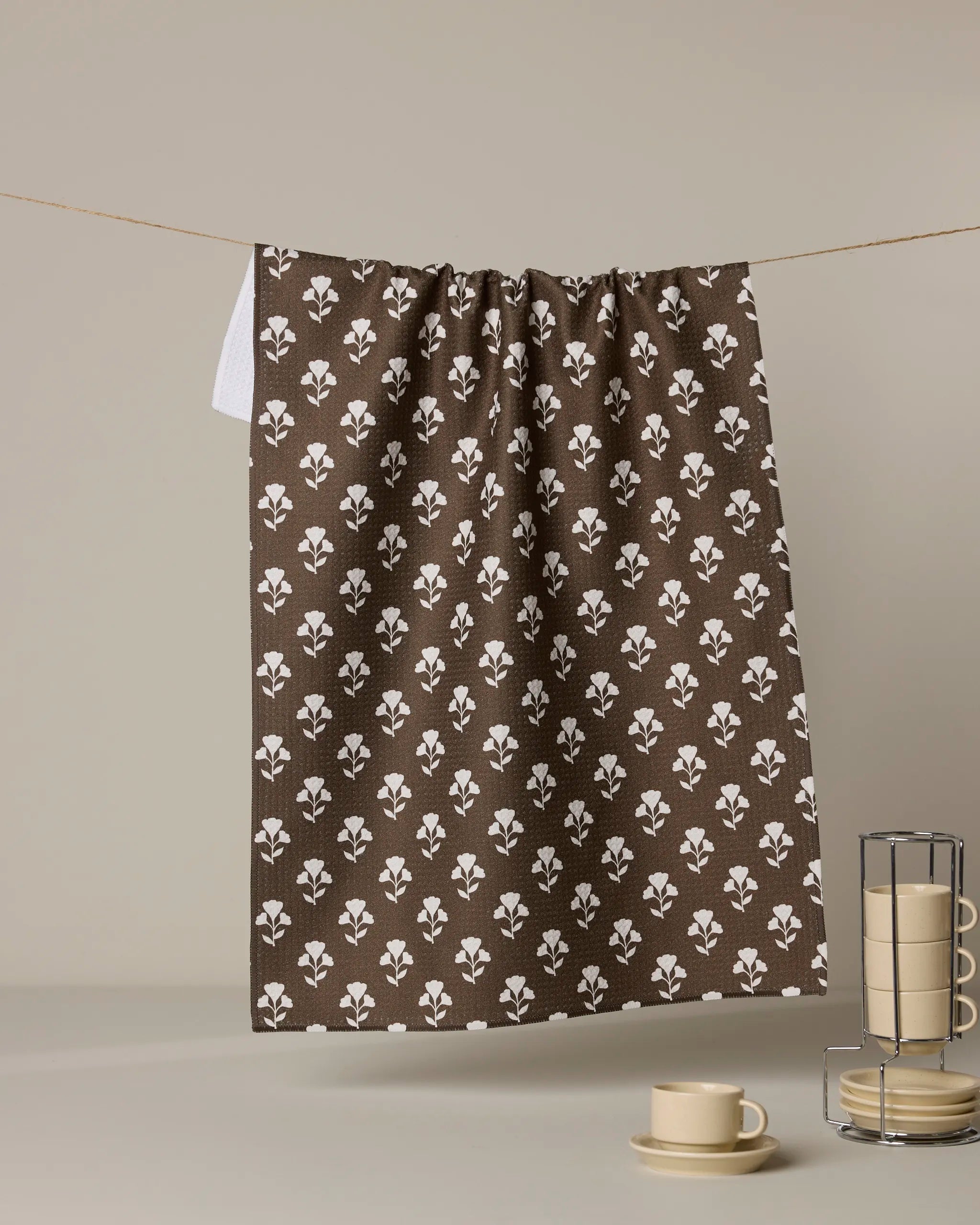 The Geometry "Bloom for Mom" ultra-absorbent brown tea towel with a white floral design hangs by a string, complemented by stacked beige cups, saucers, and plates in a metal holder against a neutral background.
