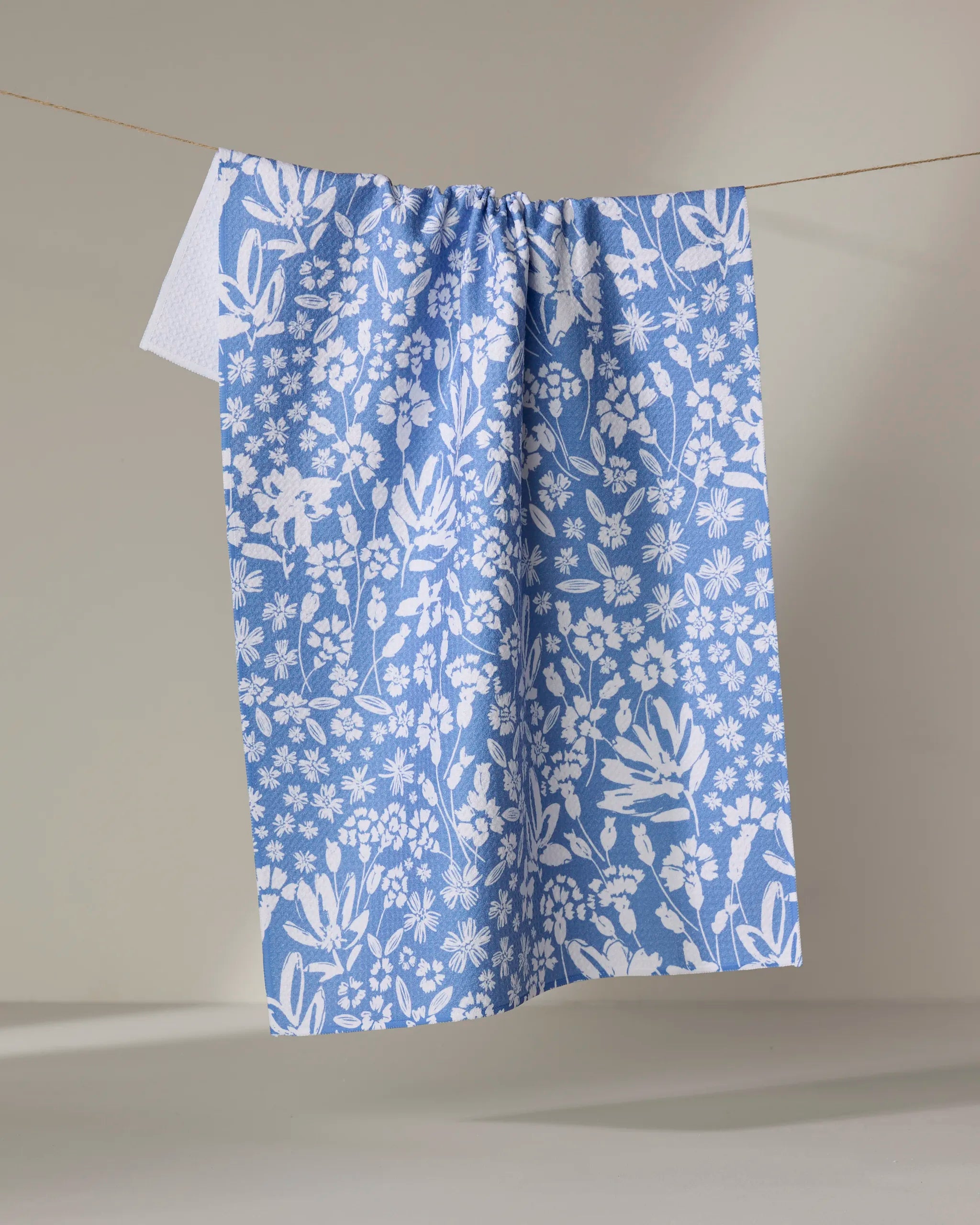 The Bloom Me Over Floral dish towel by Hufton Studio, featuring a blue and white floral pattern, hangs absorbently on a clothesline against a simple light backdrop.