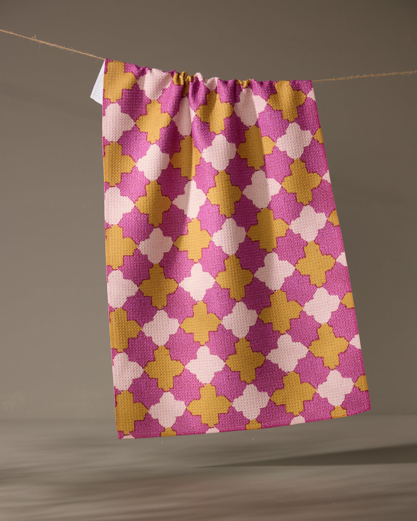 The Geometry Bloomgrid kitchen tea towel, featuring a pink, yellow, and white geometric pattern, hangs from a string against a plain neutral background.