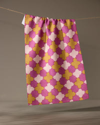 The Geometry Bloomgrid kitchen tea towel, featuring a pink, yellow, and white geometric pattern, hangs from a string against a plain neutral background.