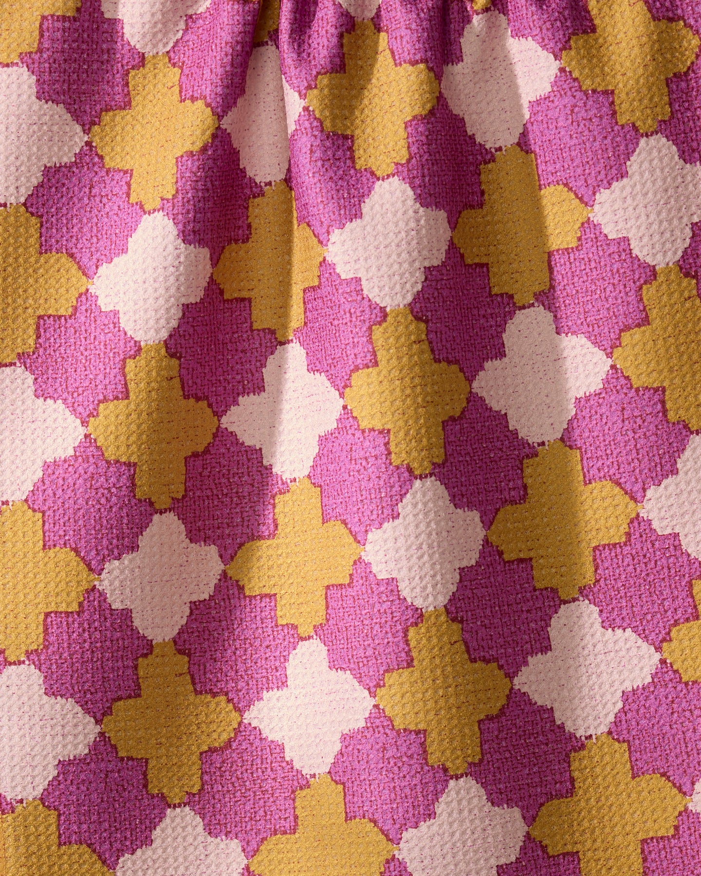 Close-up of the Geometry Bloomgrid kitchen tea towel, made from recycled materials with absorbent textured fabric. Features a repeating pink, mustard yellow, and off-white geometric pattern and slight gathered folds along the top edge.