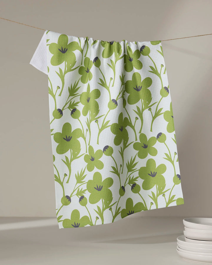 The Lizzie Clark "Blooming Blossoms" kitchen towel, featuring a green floral and leaf design on white, hangs above a stack of white plates against a neutral background.