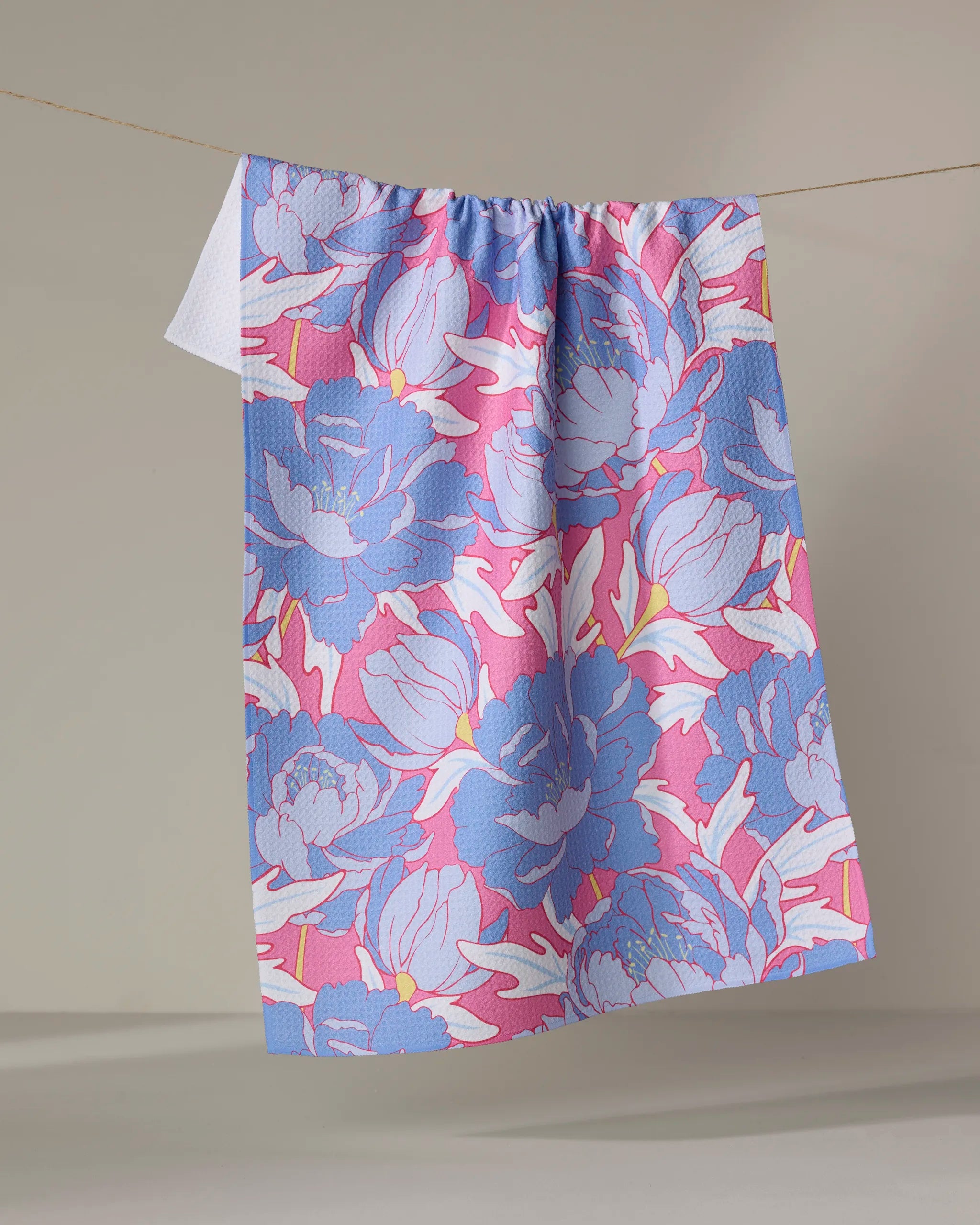 The Ashley Celena "Blooming Peonies" towel, ultra-absorbent and vibrant with blue, pink, and purple floral patterns, hangs on a clothesline against a neutral background.