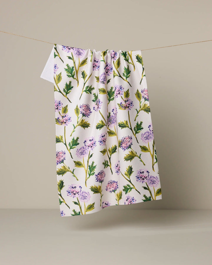 The Geometry Blooming Thistle Kitchen Tea Towel, made from recycled materials, showcases a vibrant floral pattern with green leaves and purple flowers. It hangs against a neutral beige background.
