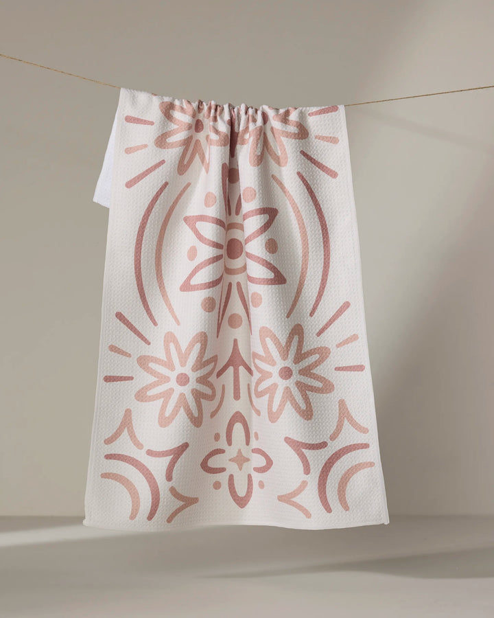 The Blossom tea towel by Adam Nienow, featuring a pink geometric floral pattern on white Geoweave fabric, hangs on a clothesline against a plain, light background.