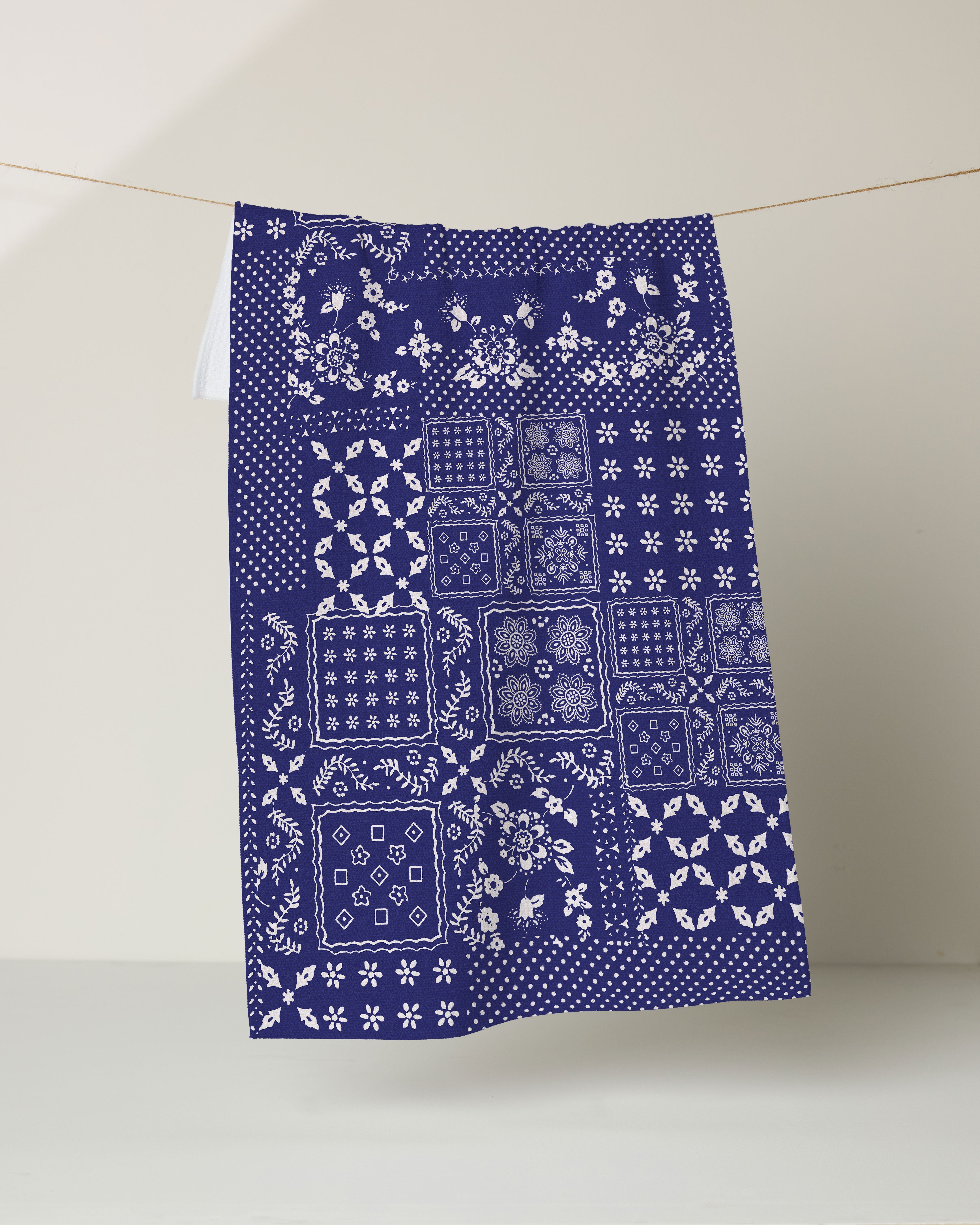 The Geometry Blossoming Bandanna Blue kitchen tea towel, crafted from recycled materials, features white floral and geometric patchwork on dark blue and hangs against a light background.