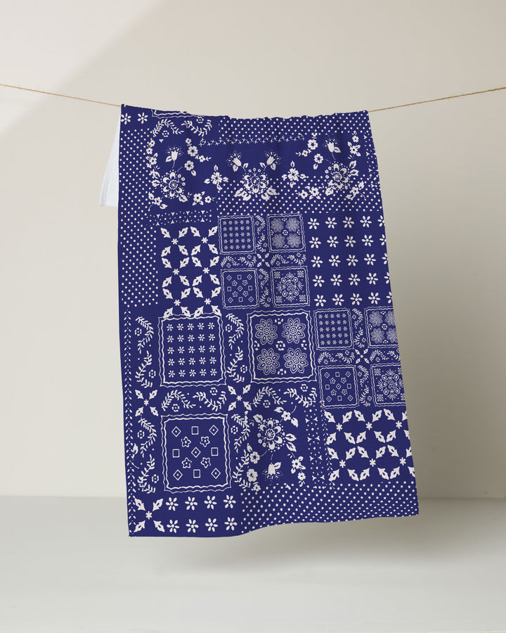 The Geometry Blossoming Bandanna Blue kitchen tea towel, crafted from recycled materials, features white floral and geometric patchwork on dark blue and hangs against a light background.