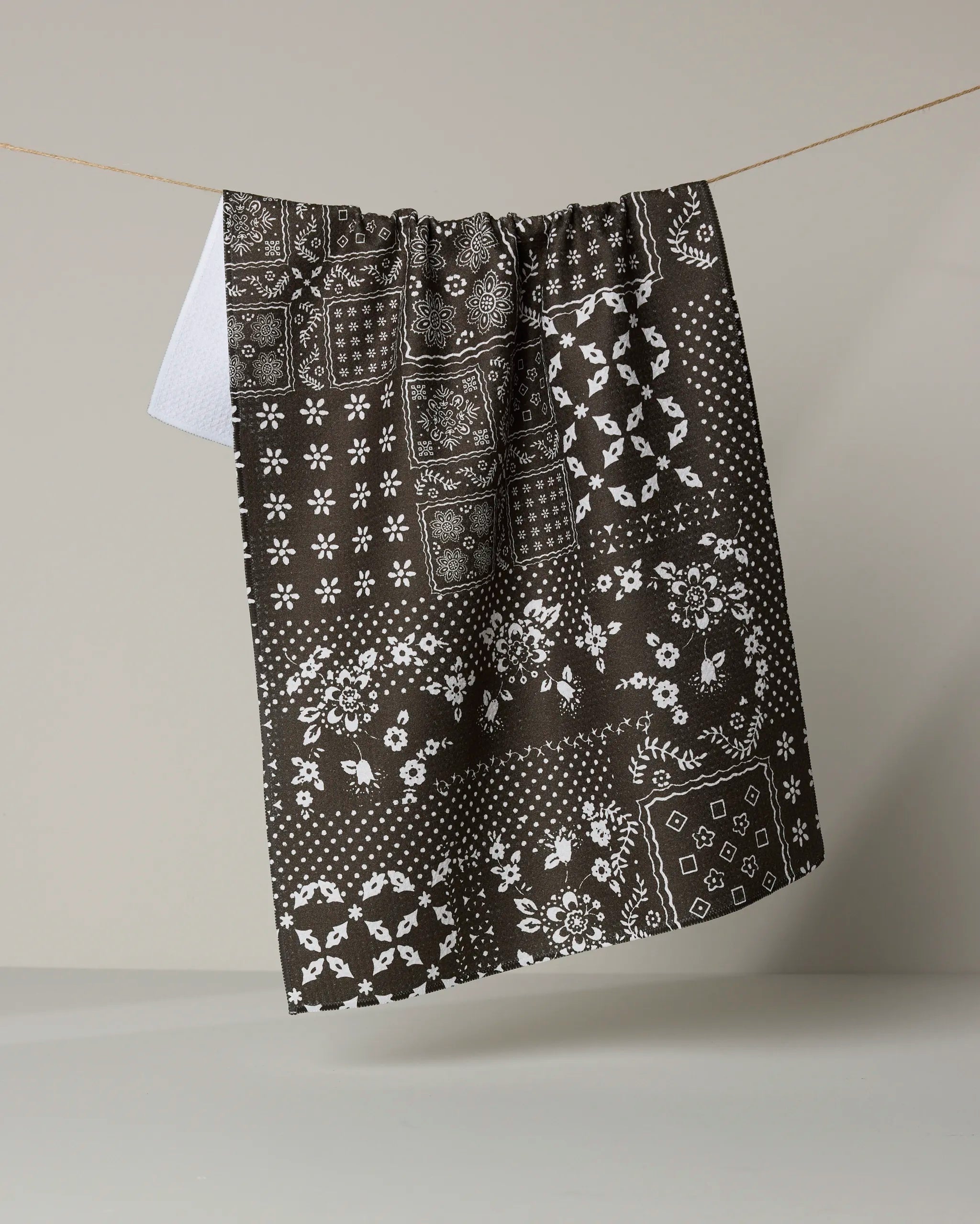 The Geometry Blossoming Bandanna Brown is an eco-friendly kitchen towel featuring a white floral and geometric bandana-style pattern, displayed hanging against a neutral background.