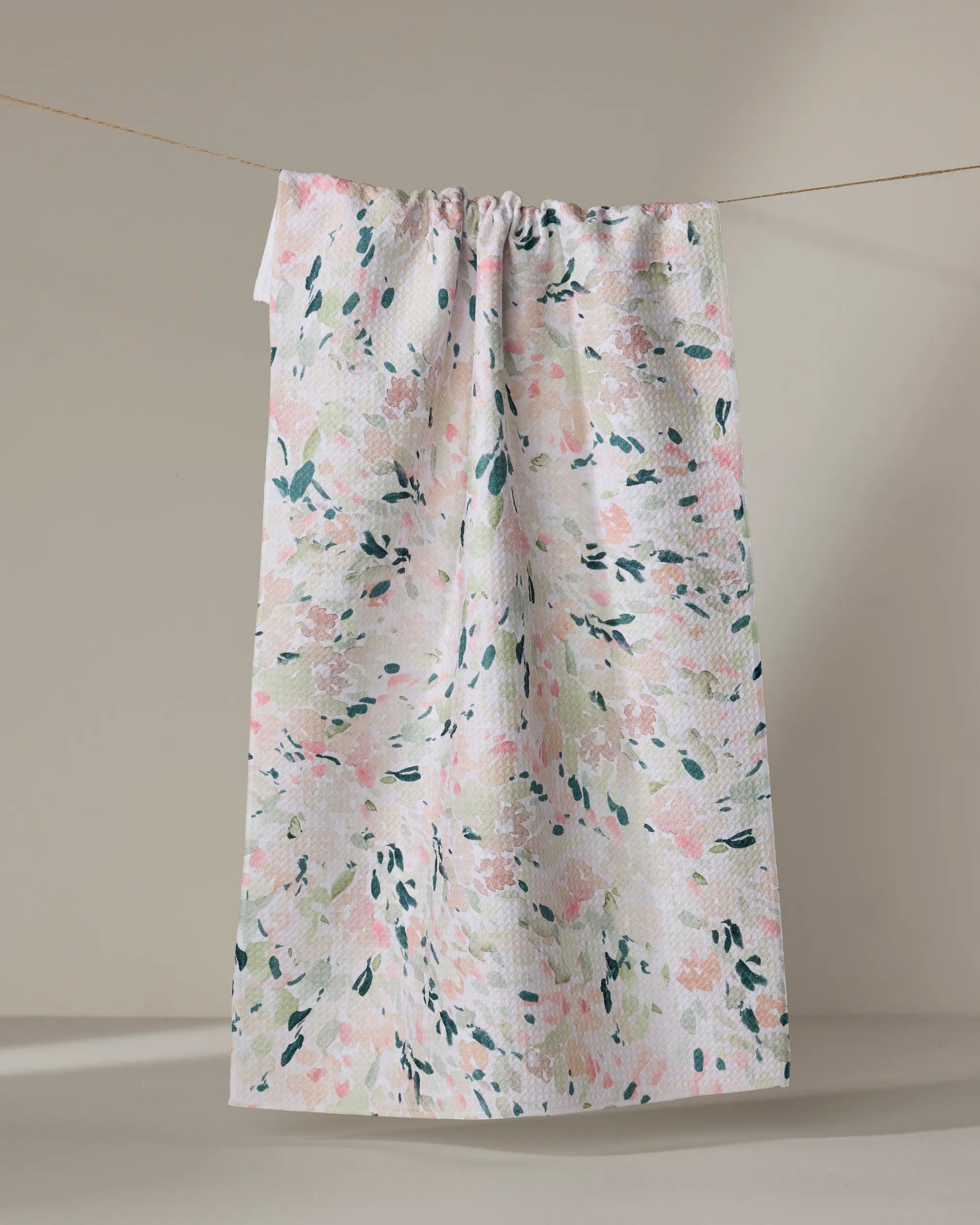 The Blossoms tea towel by Anee Shah features a soft pastel floral pattern in pink, green, and cream. Made from ultra-absorbent recycled materials, it blends eco-friendly style and function, shown hanging against a neutral background.