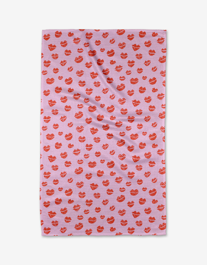 The Kayla Ann Blowing Kisses tea towel is pink and rectangular, featuring a red lips pattern. Made from recycled materials, it’s shown laid flat with slight wrinkles visible.