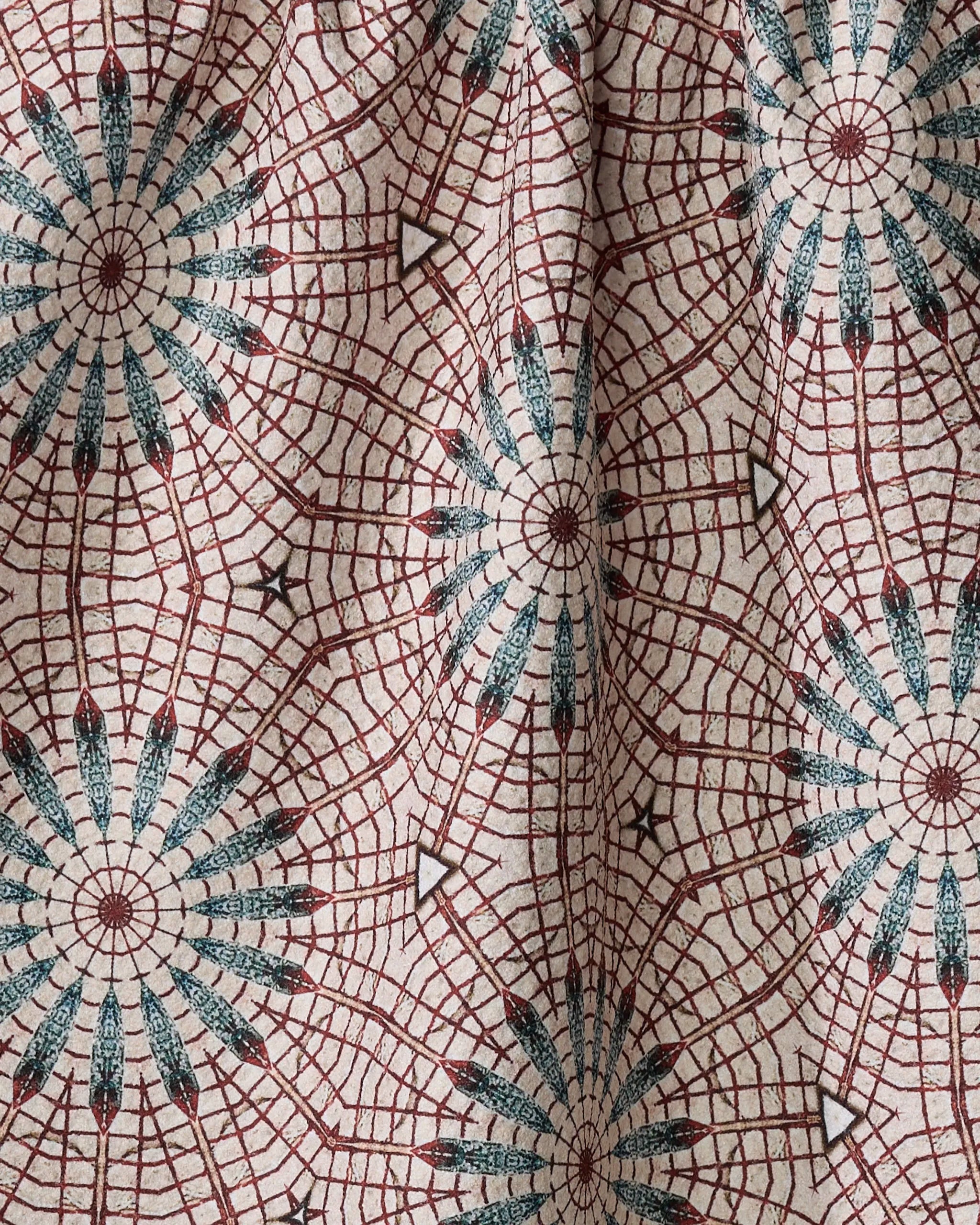 A close-up of the Blue Aster Foxtrot kitchen towel by Katherine Berkeley, made from recycled materials with red and blue circular, web-like patterns on a light beige background. Slight wrinkles add shadows and texture.