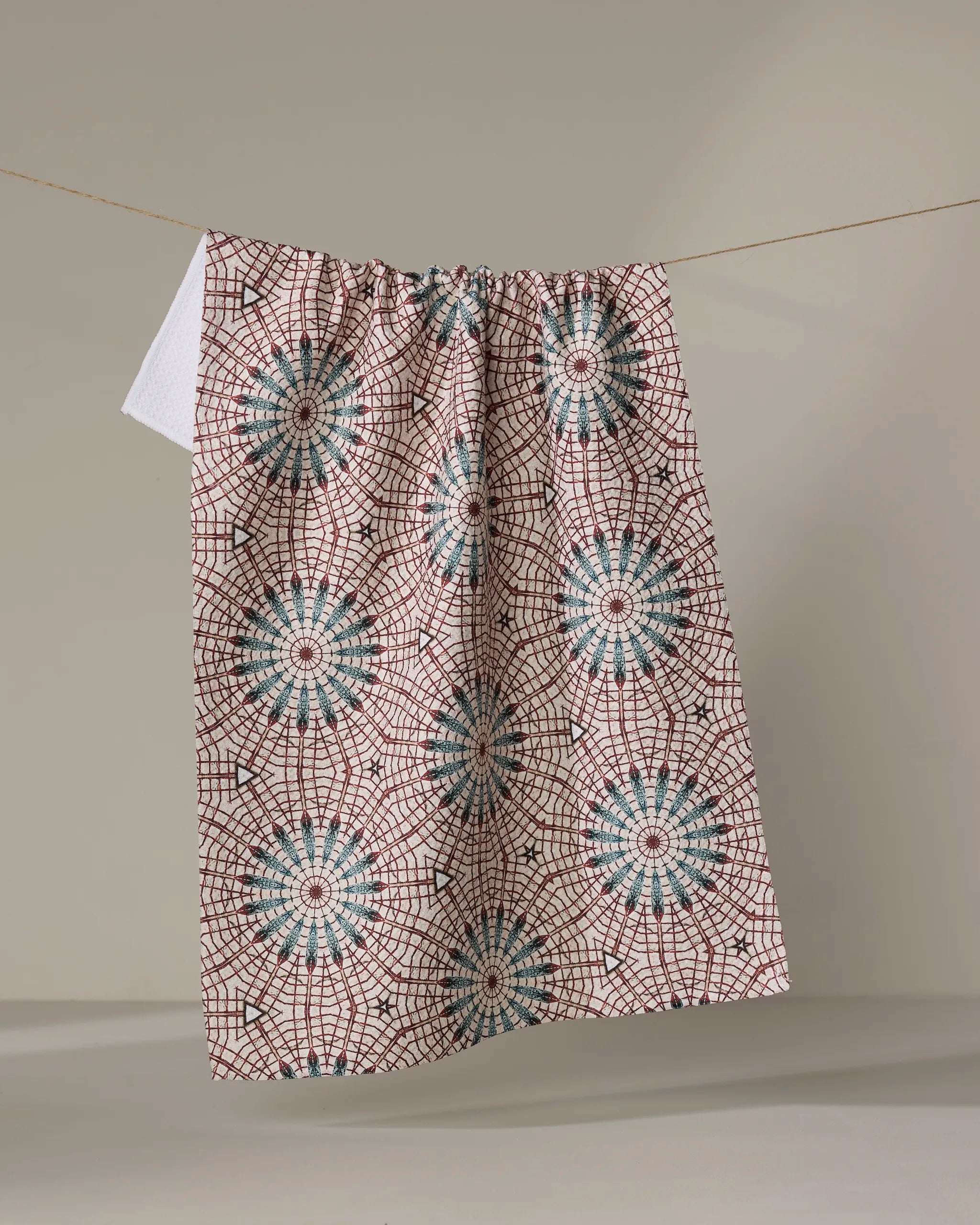 The Katherine Berkeley Blue Aster Foxtrot kitchen towel, featuring blue and red geometric starburst patterns and made from recycled materials, hangs on a clothesline against a plain, neutral background.
