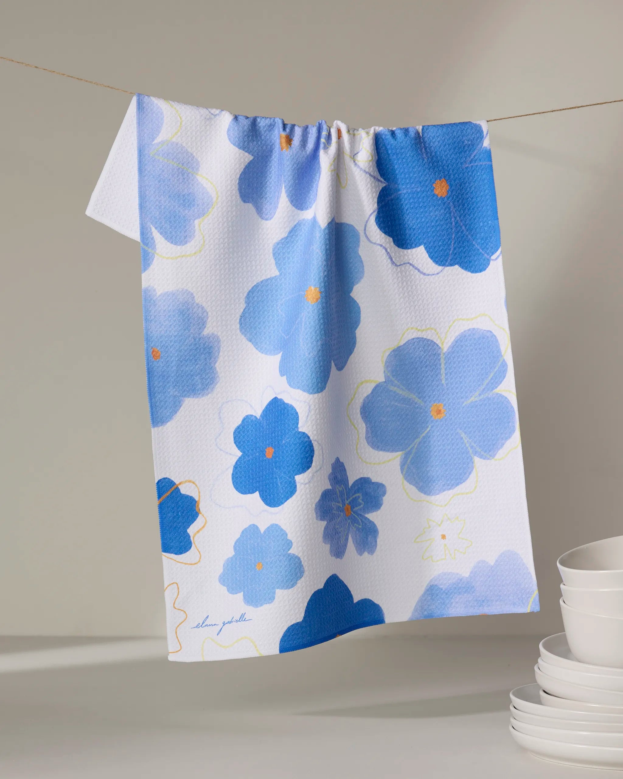 The Blue Blossoms kitchen towel by Elana Gabrielle, featuring large blue and light blue floral patterns, hangs on a line. In the corner, eco-friendly white bowls are stacked against a neutral background.