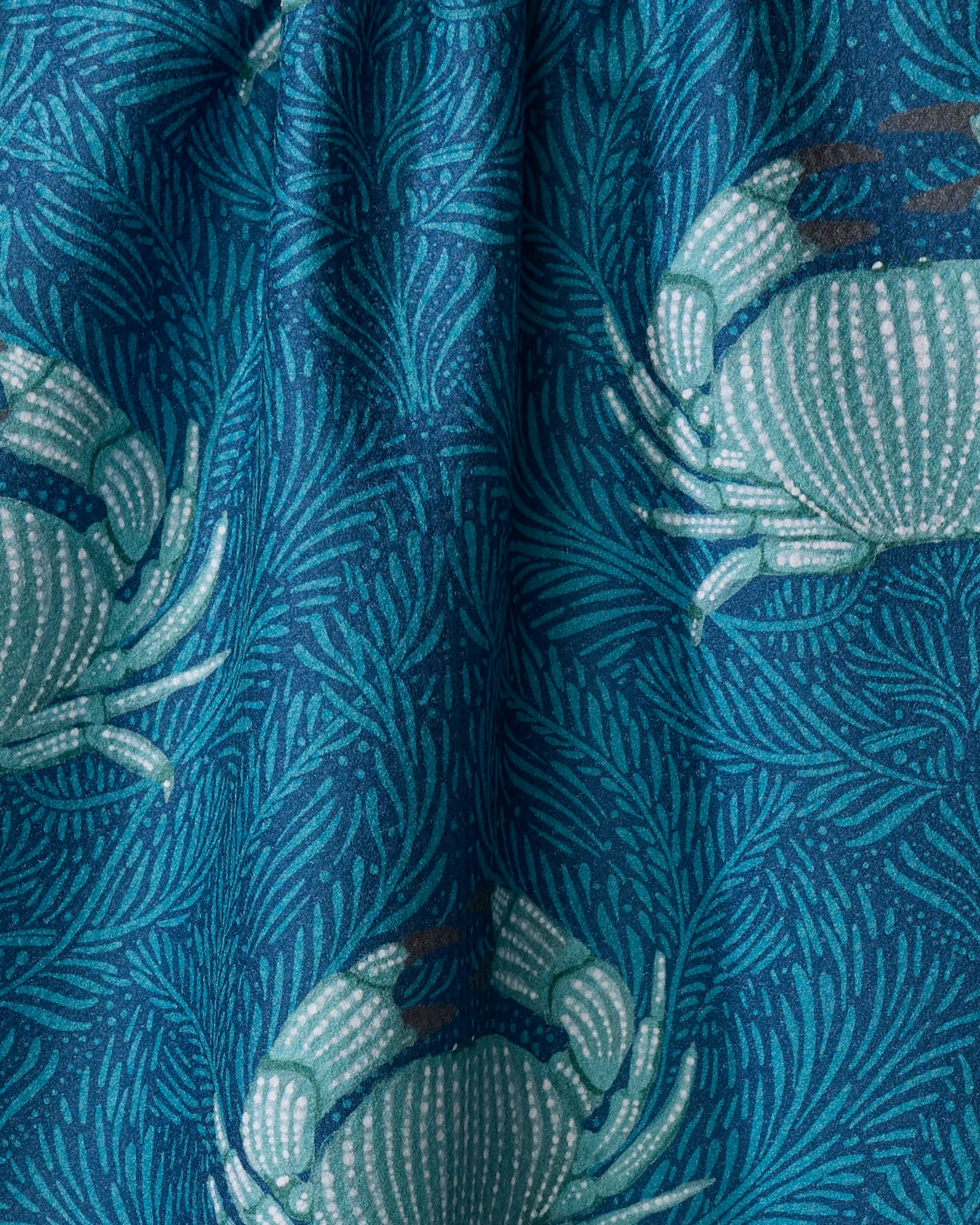 The "Blue Crab" tea towel by Katie O'Shea features a blue fabric with light blue crab and leaf-like patterns. Made from ultra-absorbent material, its slightly wrinkled texture adds depth and visual interest.