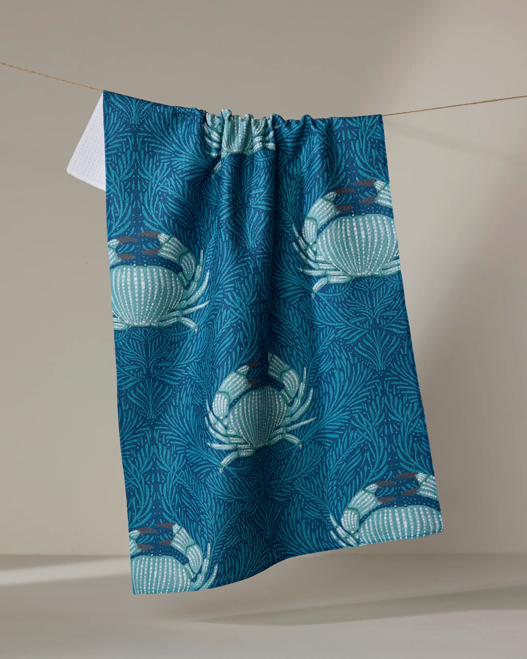 The "Blue Crab" tea towel by Katie O'Shea, made from recycled materials and featuring white crabs and seaweed on blue, hangs on a clothesline against a neutral background.