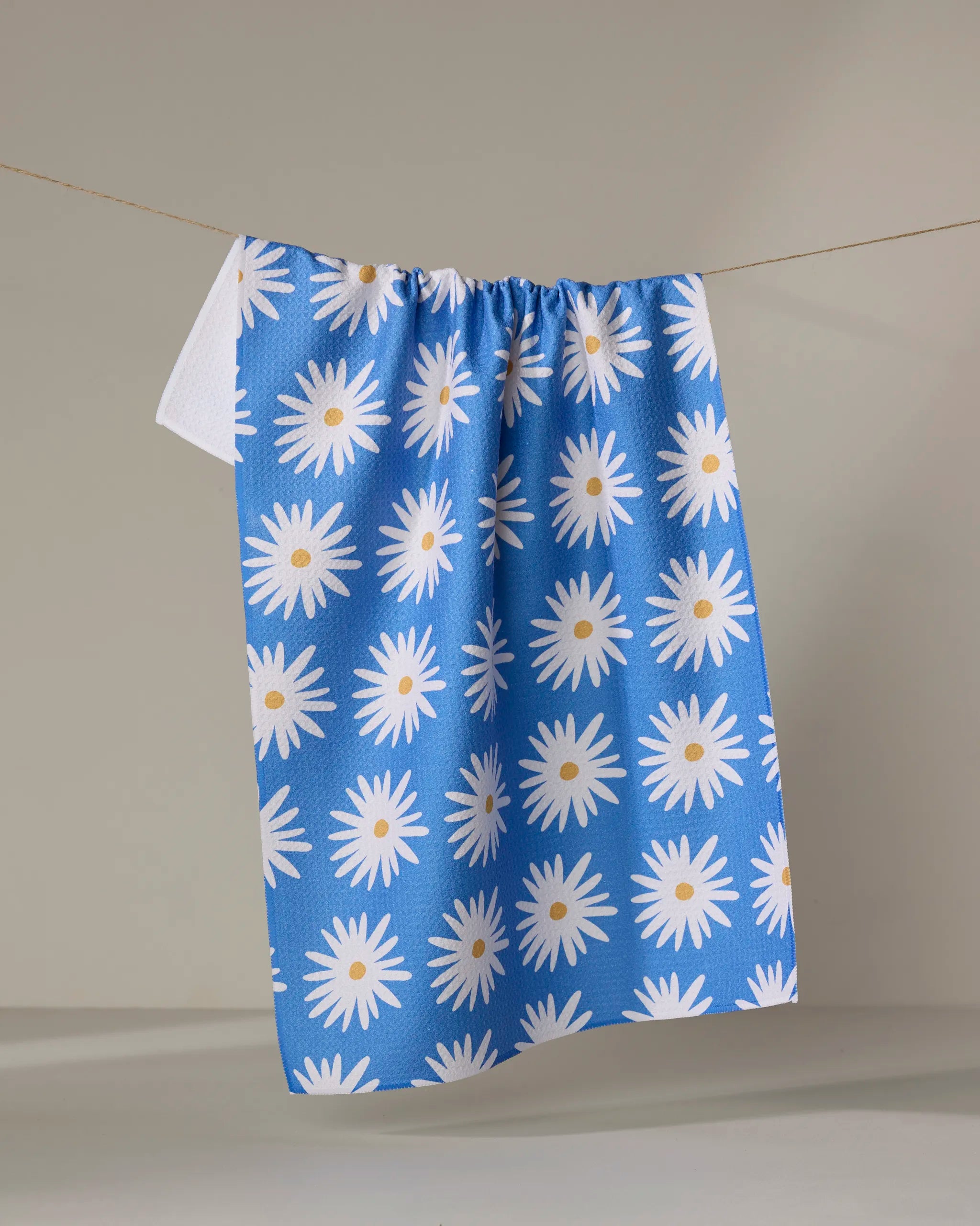 The Blue Daisies tea towel by Megan Roy, made from recycled materials and featuring a white daisy floral pattern, hangs on a clothesline against a neutral background.